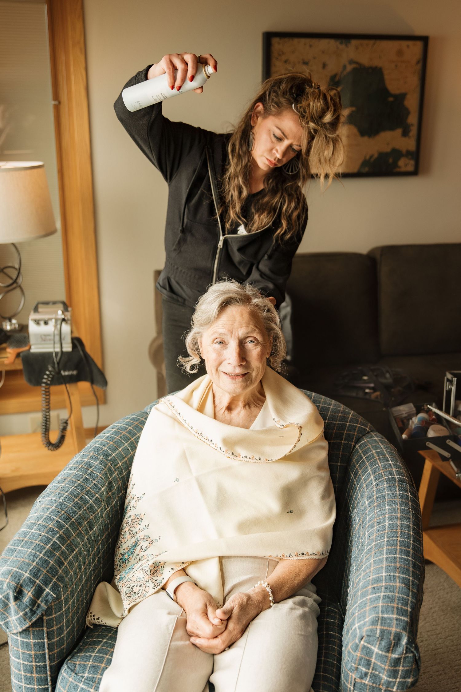 Hairstylist spraying hairspray on elderly woman seated in armchair during wedding prep.