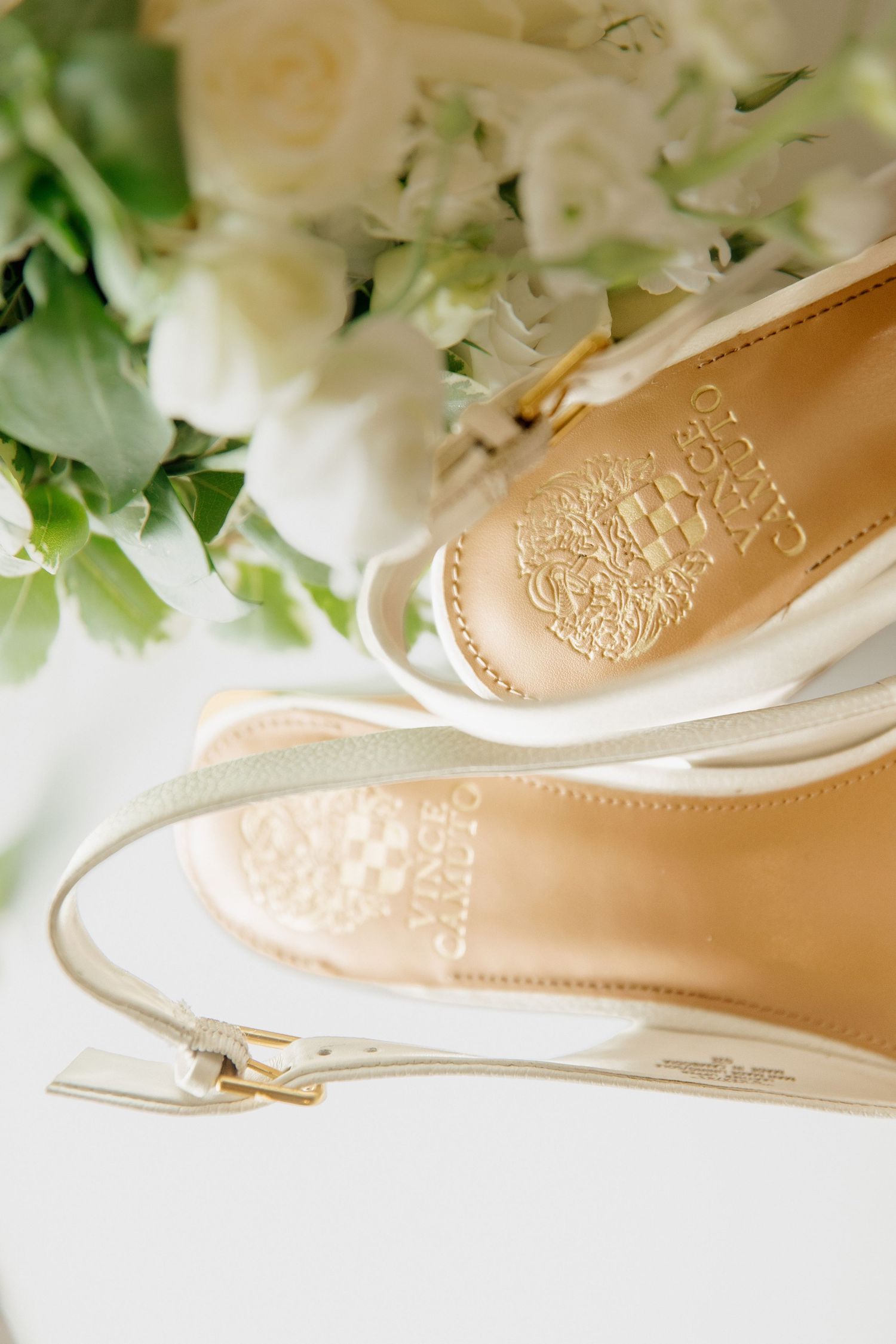 Vince Camuto bridal heels with gold insoles and white ankle straps rest beside a white floral bouquet.