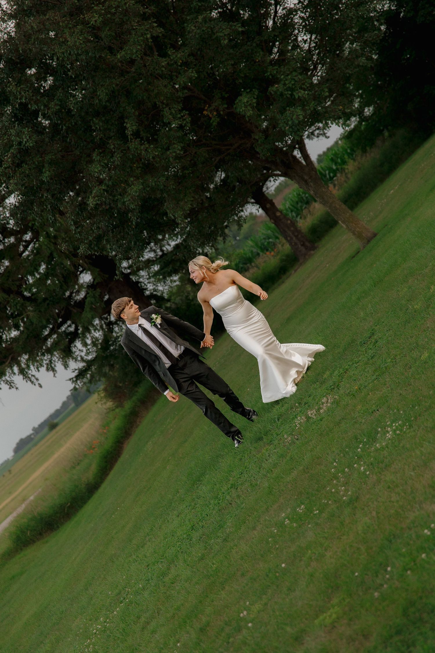 A bride in a white gown and groom in a black suit hold hands running across a lush green field under large trees.