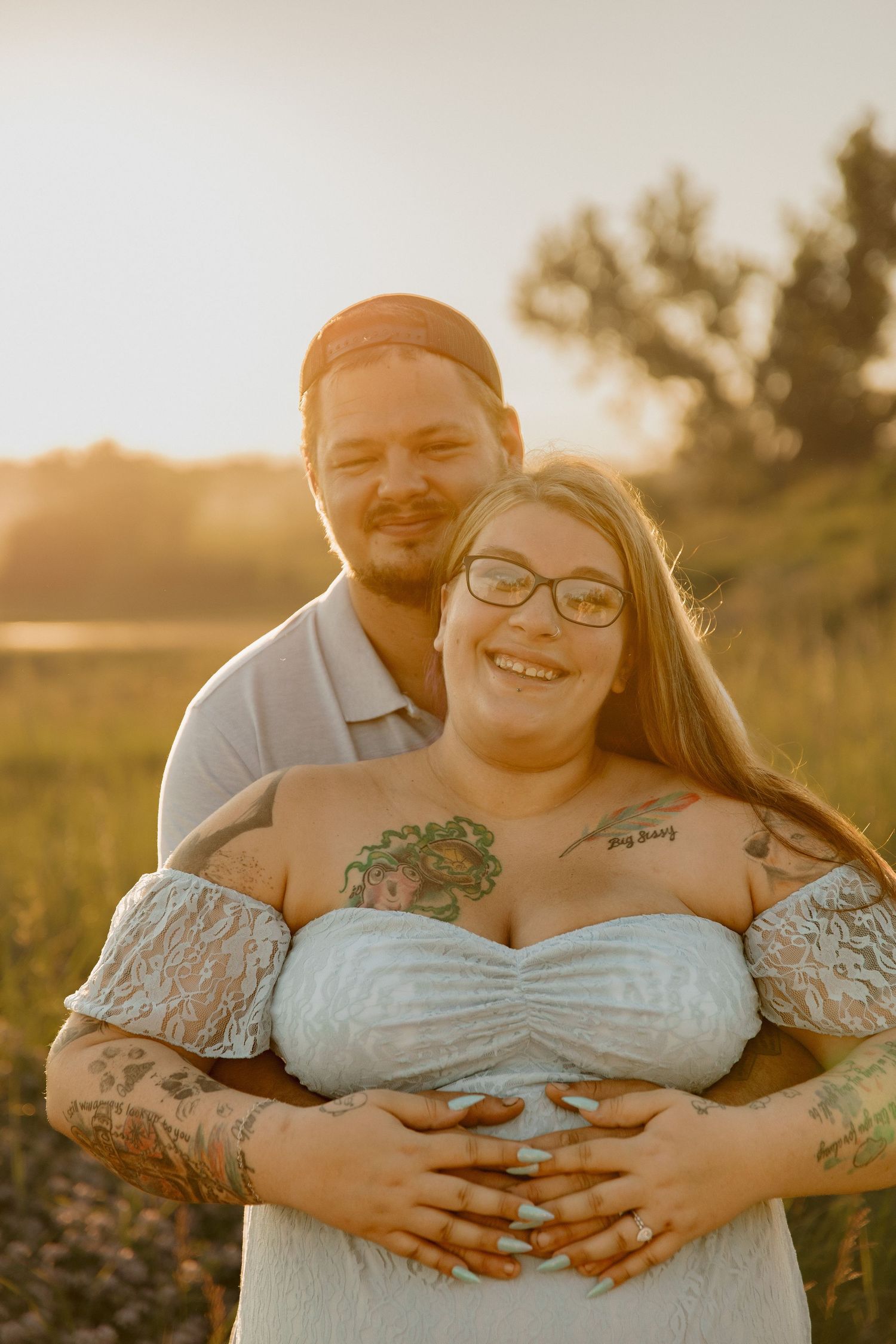 Couple embracing at golden hour sunset in a field, woman in white lace dress with tattoos, man smiling warmly behind her.