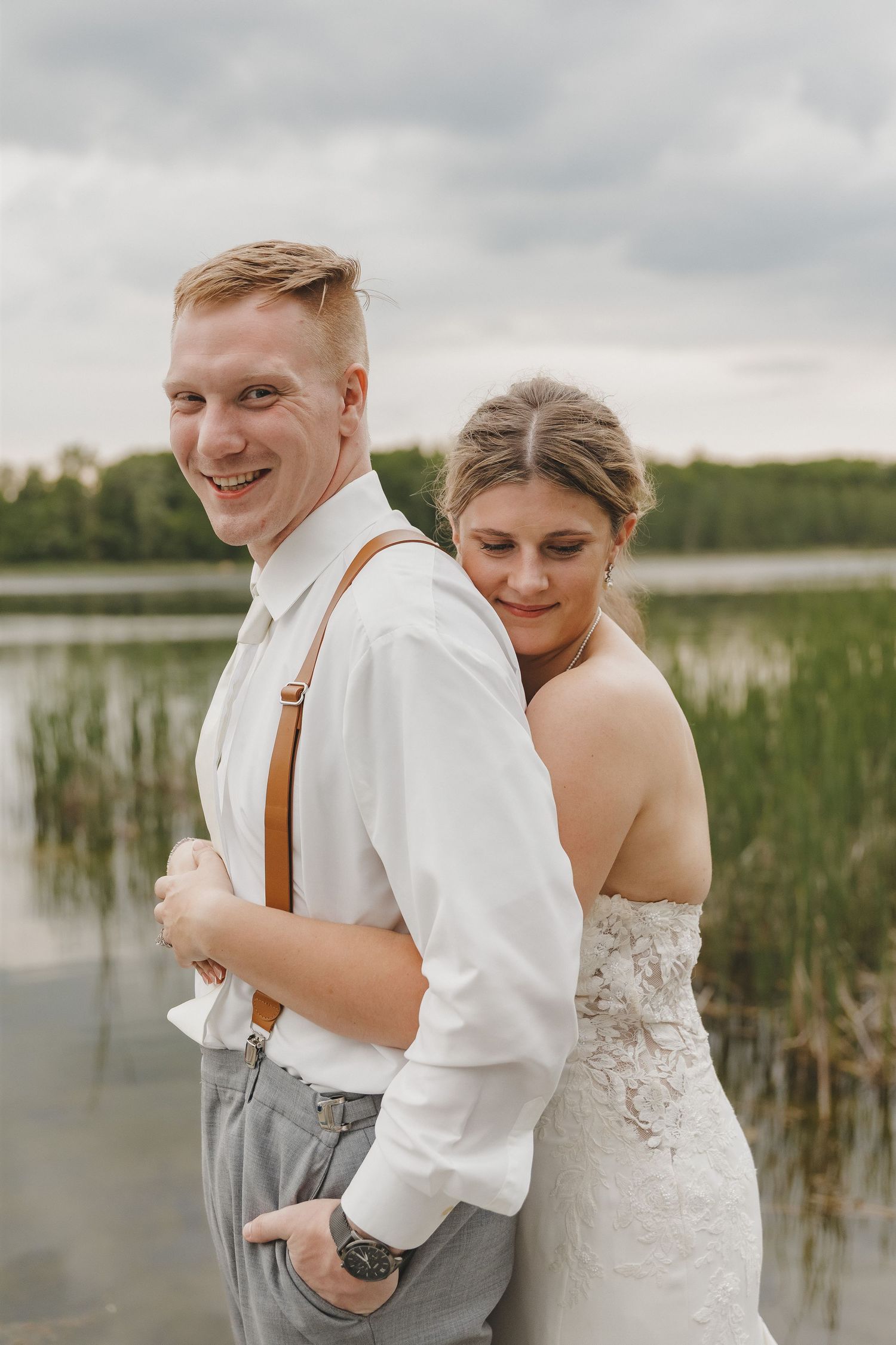 Smiling groom in white shirt and suspenders with bride hugging him from behind near a scenic lake.
