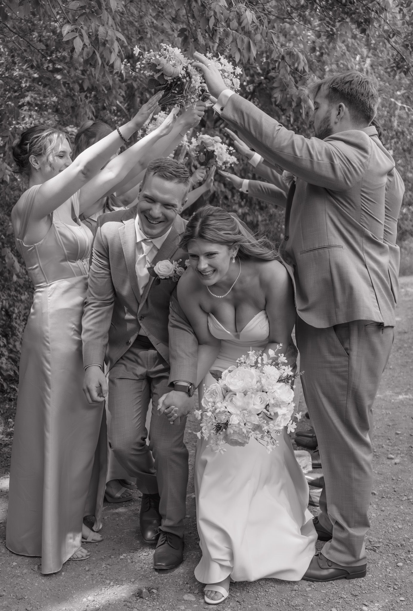 Black and white photo of smiling bride and groom walking through arch formed by wedding party.