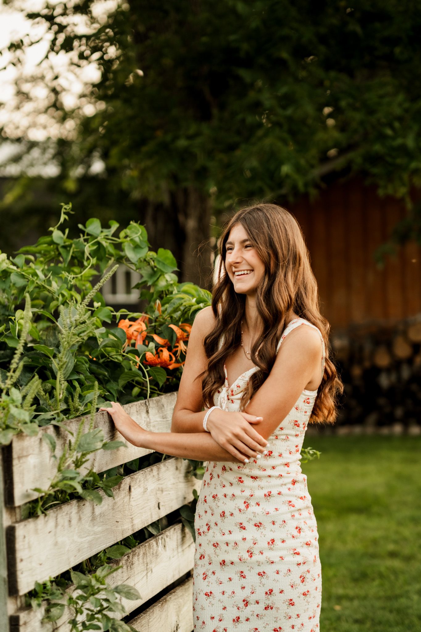 Woman in floral dress poses by rustic white fence in summer garden.