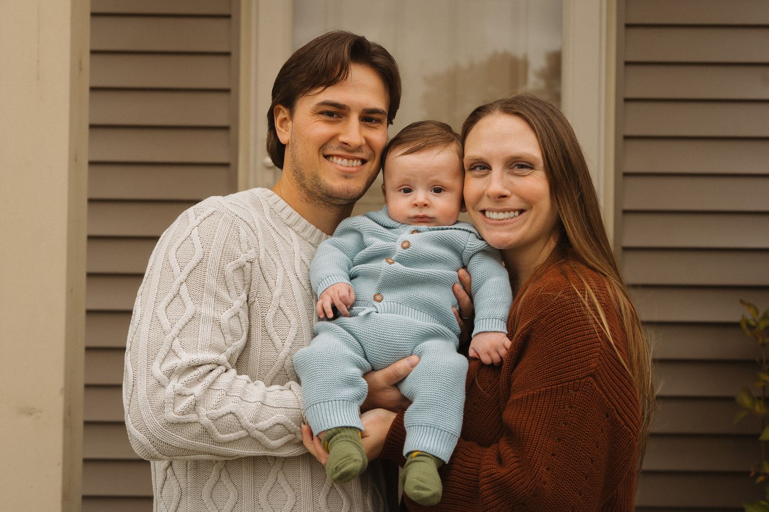 A family of three poses together for a portrait in front of beige shutters.