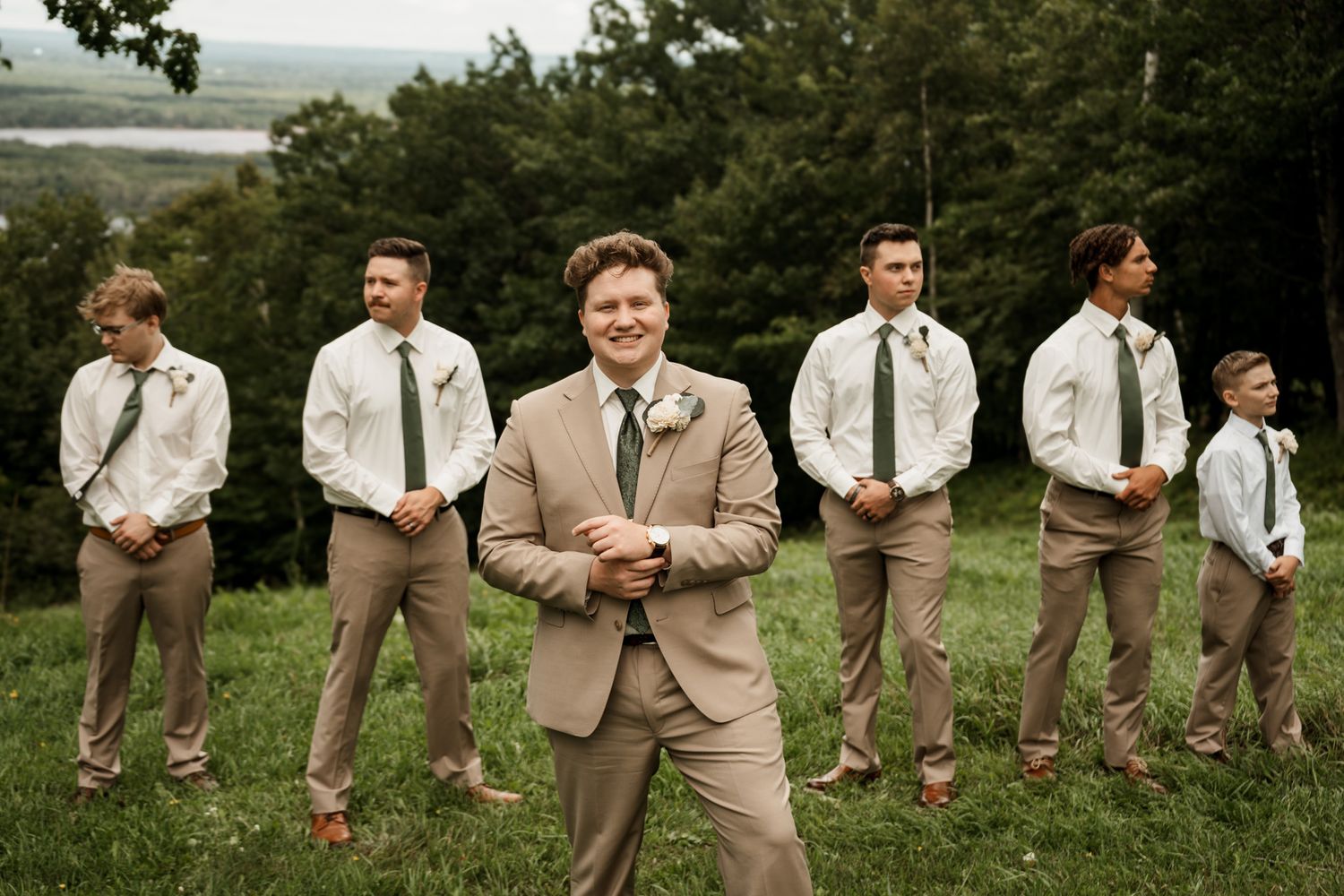 Groomsmen in tan suits pose together on a hilltop overlooking a scenic landscape.