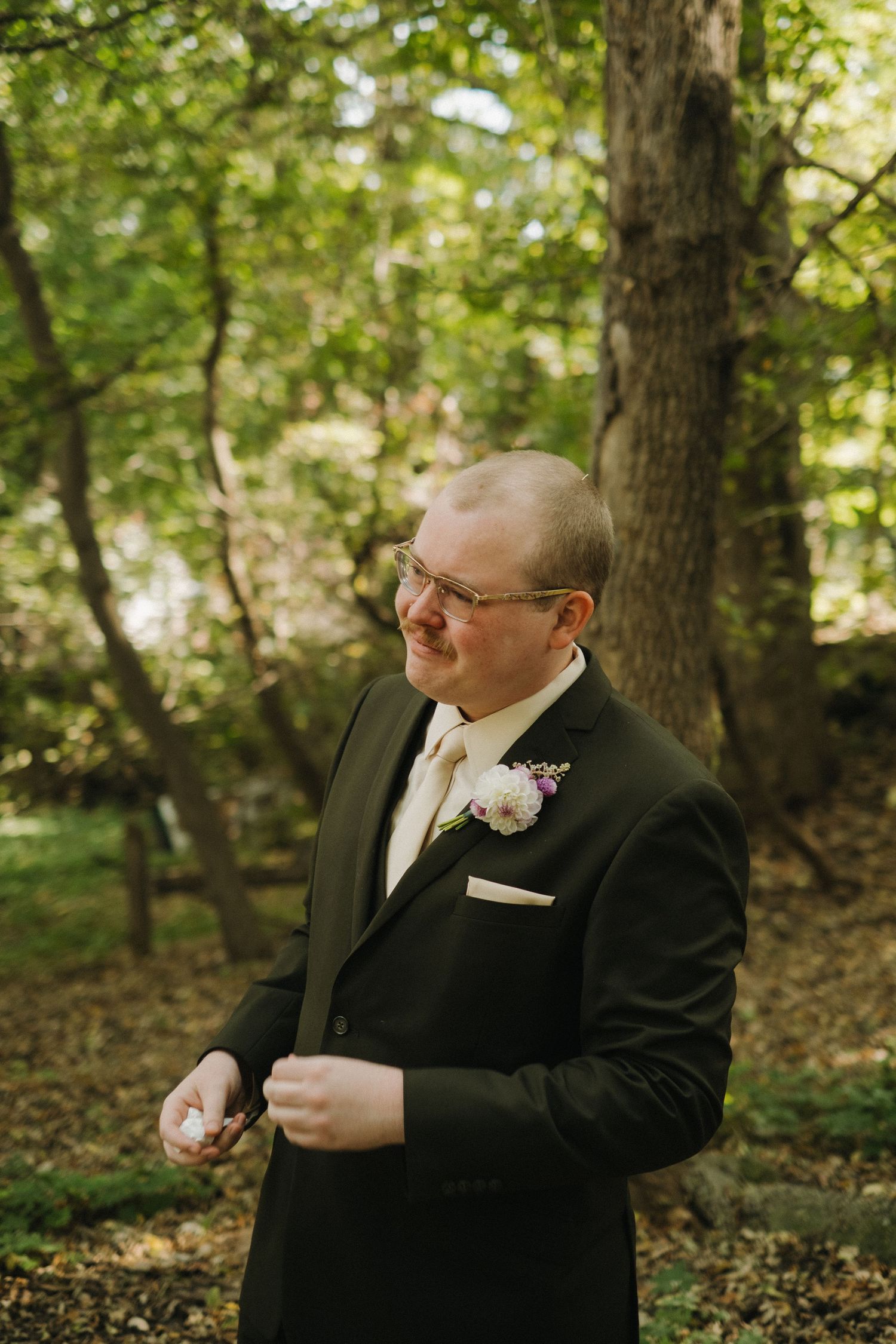 A groom in formal attire stands among trees during an outdoor wedding.