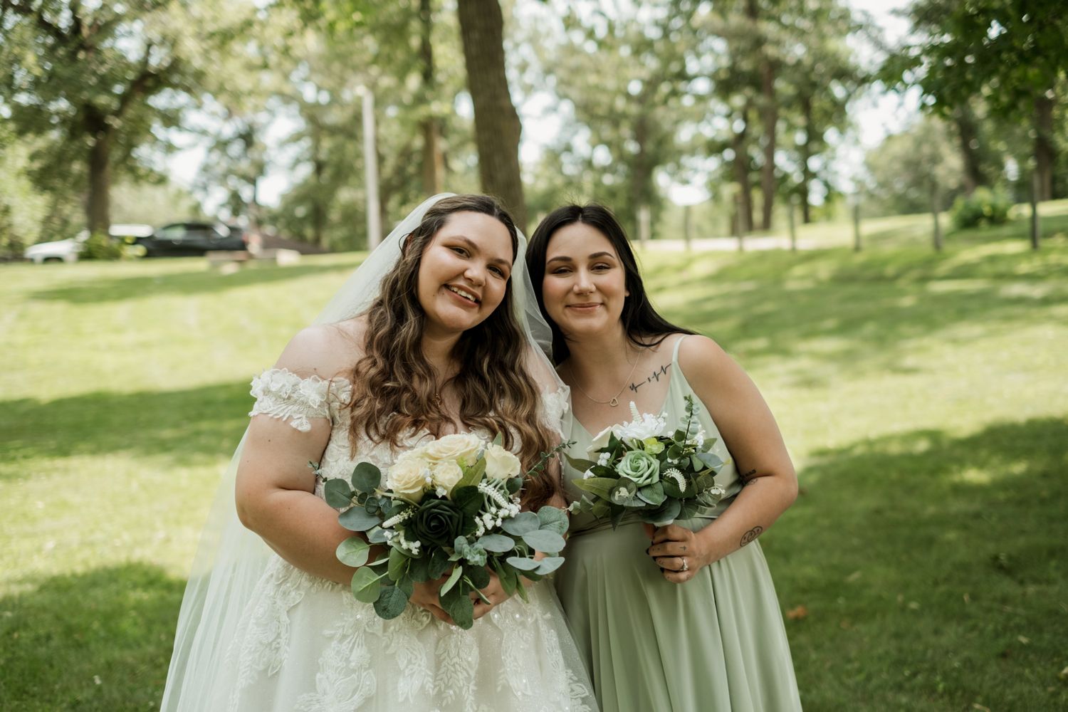 A bride and bridesmaid holding bouquets pose together in a sunny outdoor setting.