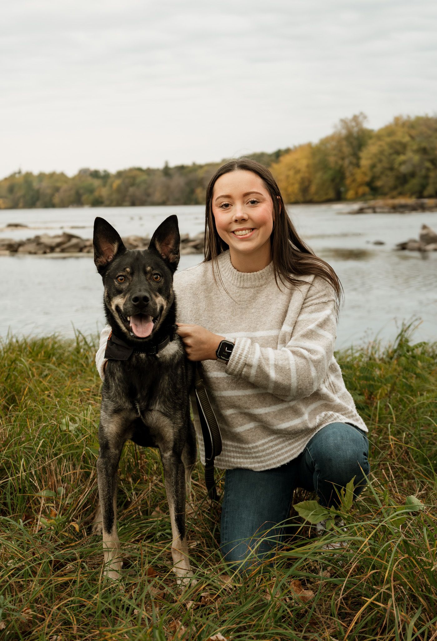 Person in cream sweater sitting with black dog by a lake in autumn.