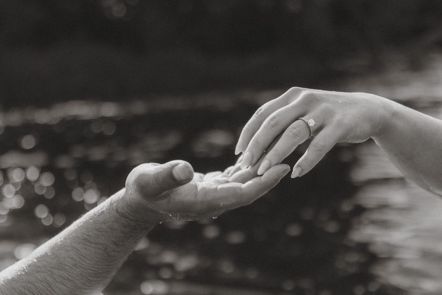Black and white photo of two hands gently touching, one wearing an engagement ring, with bokeh water background.