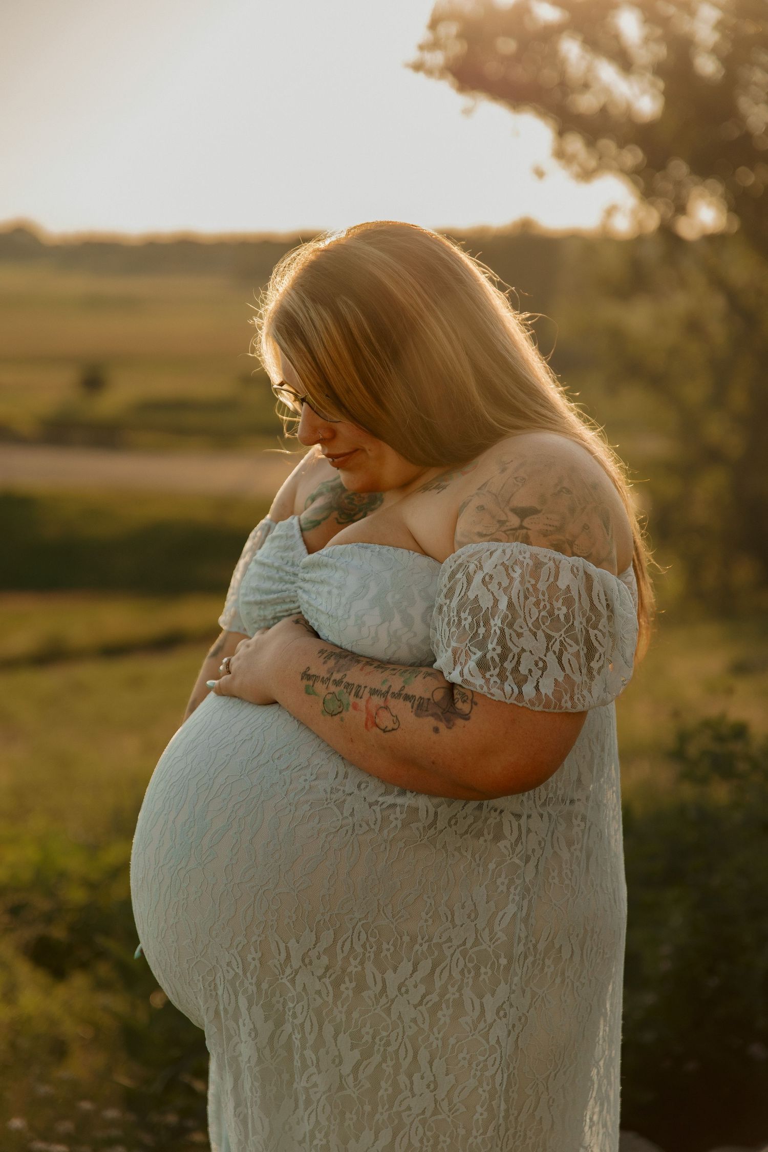 Pregnant woman in white lace off-shoulder dress cradling baby bump at golden hour in a green field.