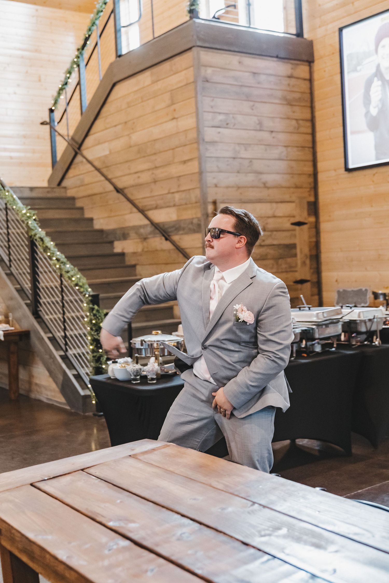 Groom in gray suit and sunglasses poses confidently inside rustic wooden wedding reception venue.