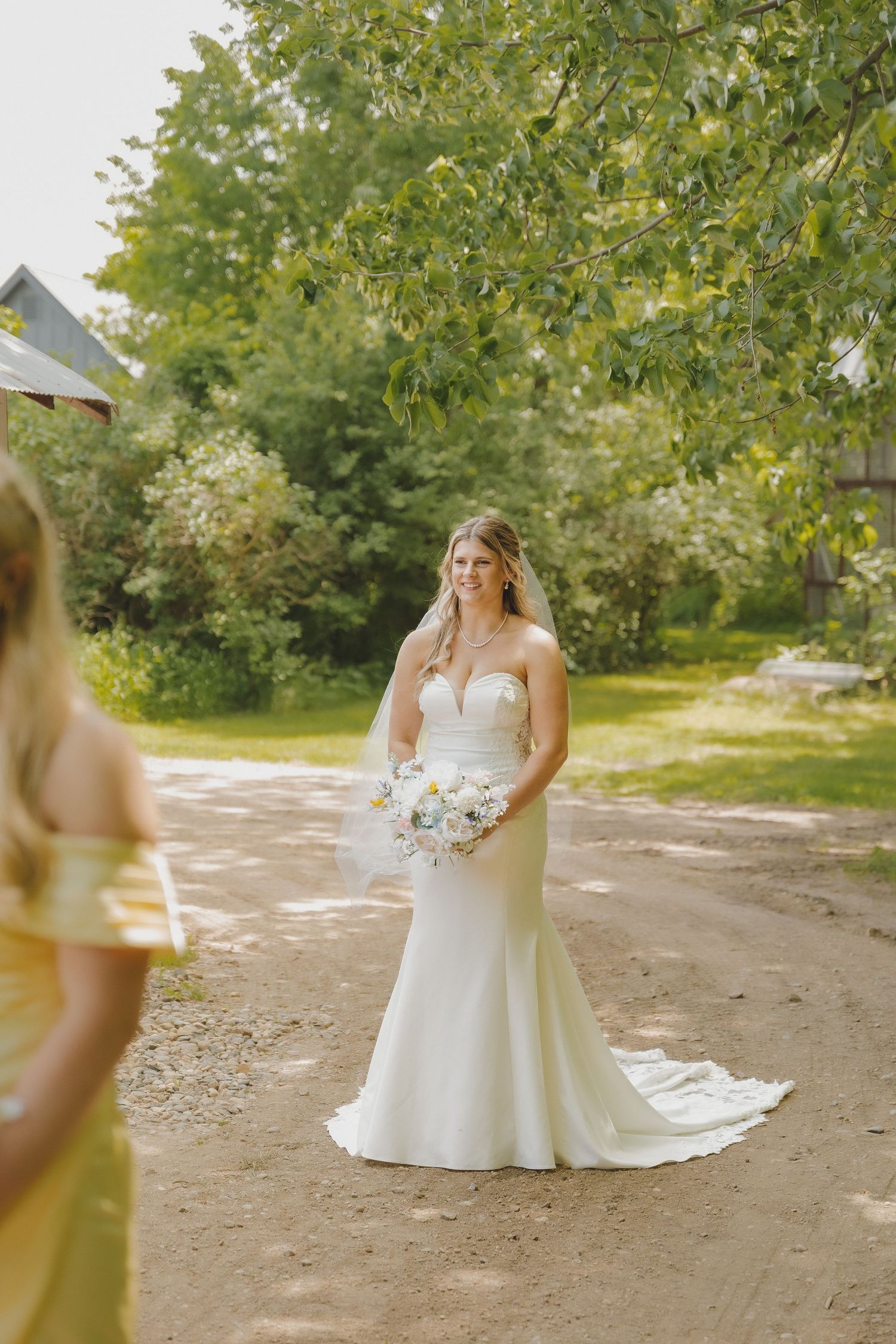 Smiling bride in strapless white mermaid gown holding white bouquet stands on dirt path with green trees behind her.