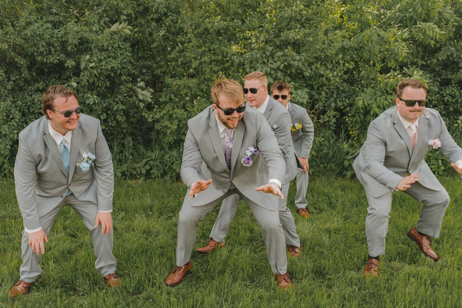 Groomsmen in gray suits and sunglasses strike fun dance poses outdoors with green trees in the background.