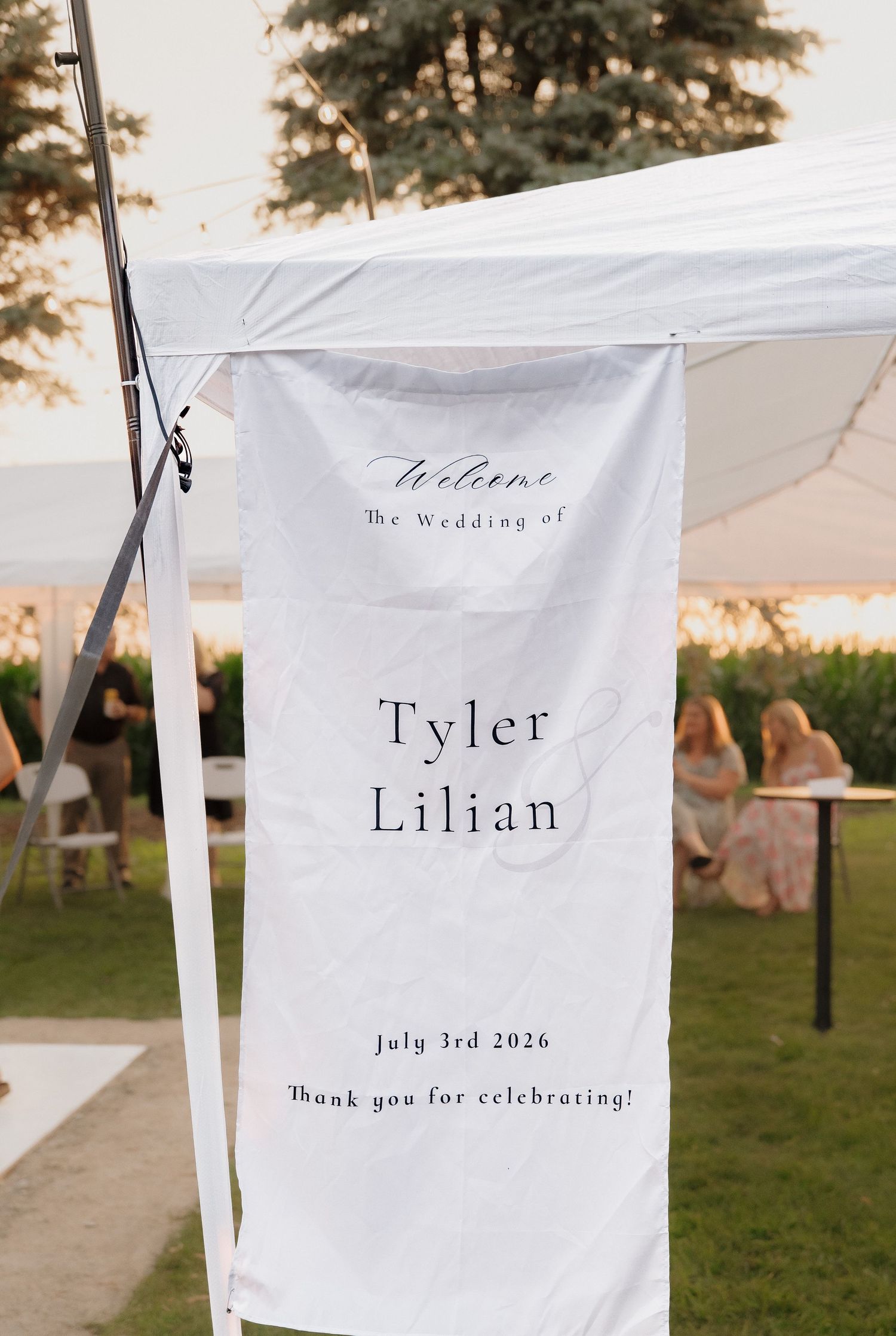 Wedding welcome banner for Tyler and Lilian, July 3rd 2026, hanging on a white tent at an outdoor reception.