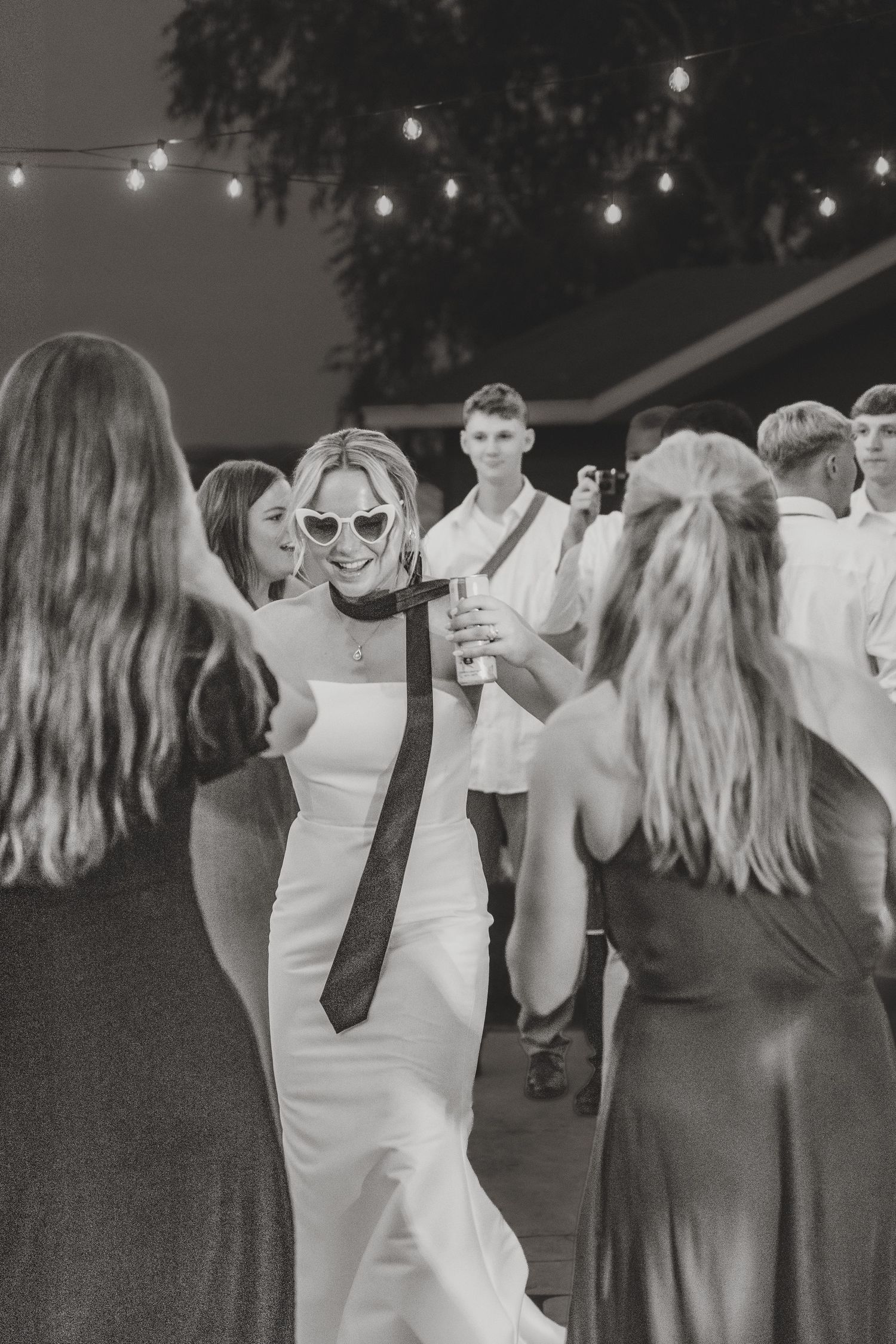 Black and white photo of bride in heart sunglasses dancing with guests at outdoor wedding reception.
