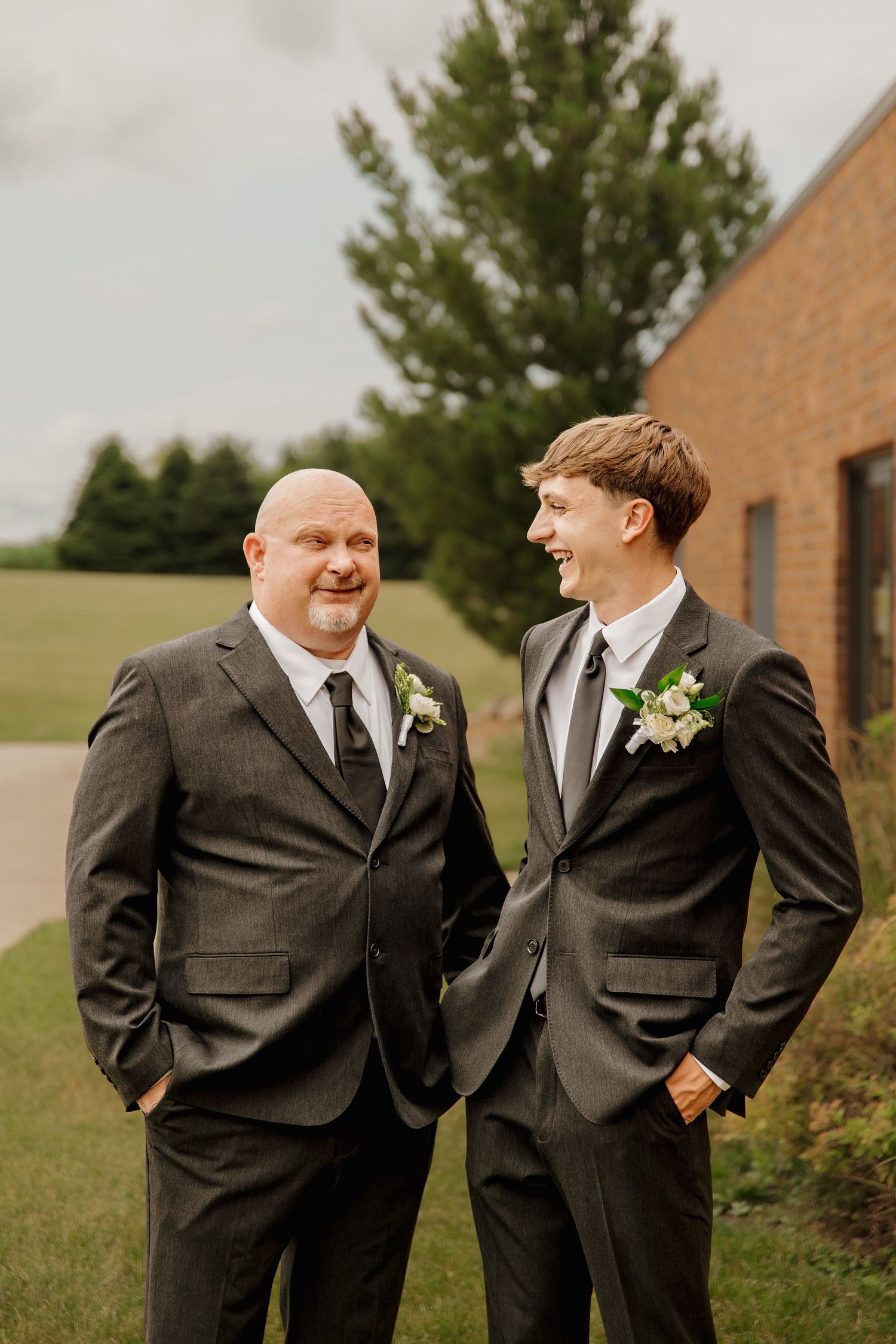 Two groomsmen in dark suits with boutonnieres smiling together outdoors near brick building.