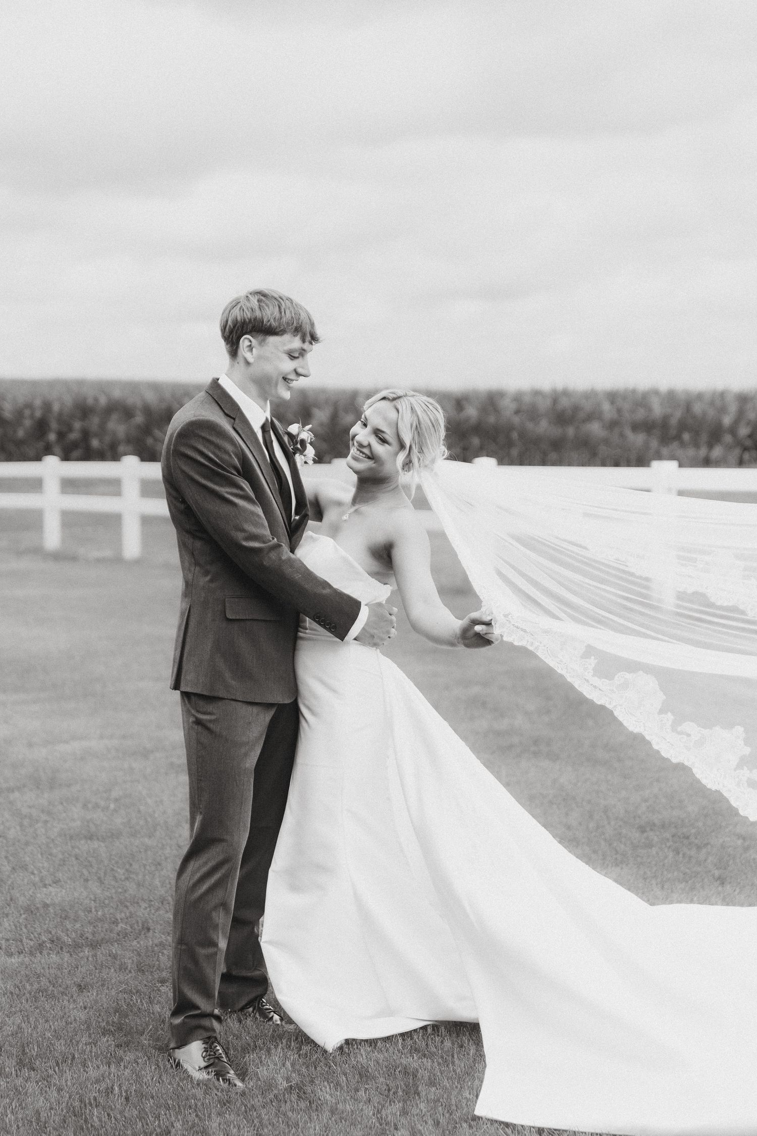 Bride and groom laughing together outdoors as her lace veil blows in the wind in black and white photo.