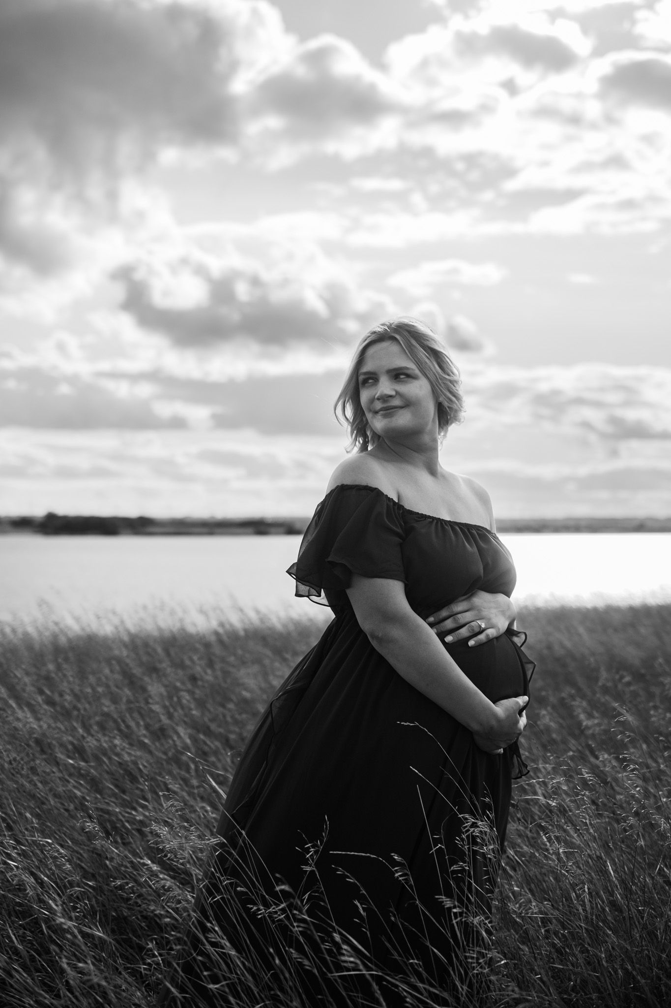 Black and white maternity photo of pregnant woman in off-shoulder dress standing in field near water under cloudy sky.