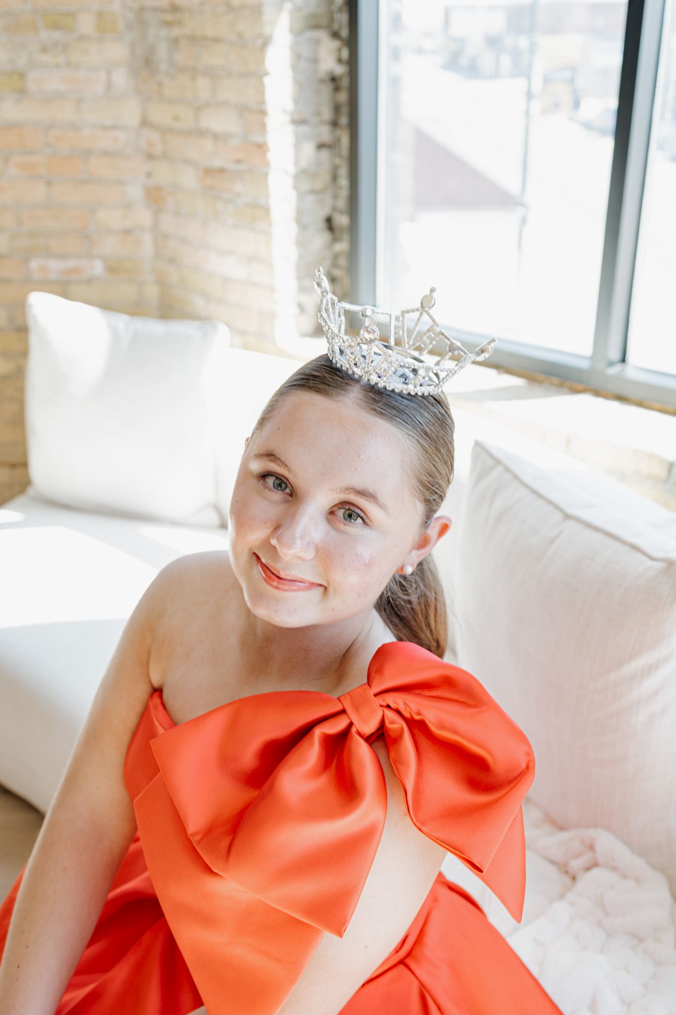 Young woman in orange bow dress with crystal crown sitting in bright windowed studio.