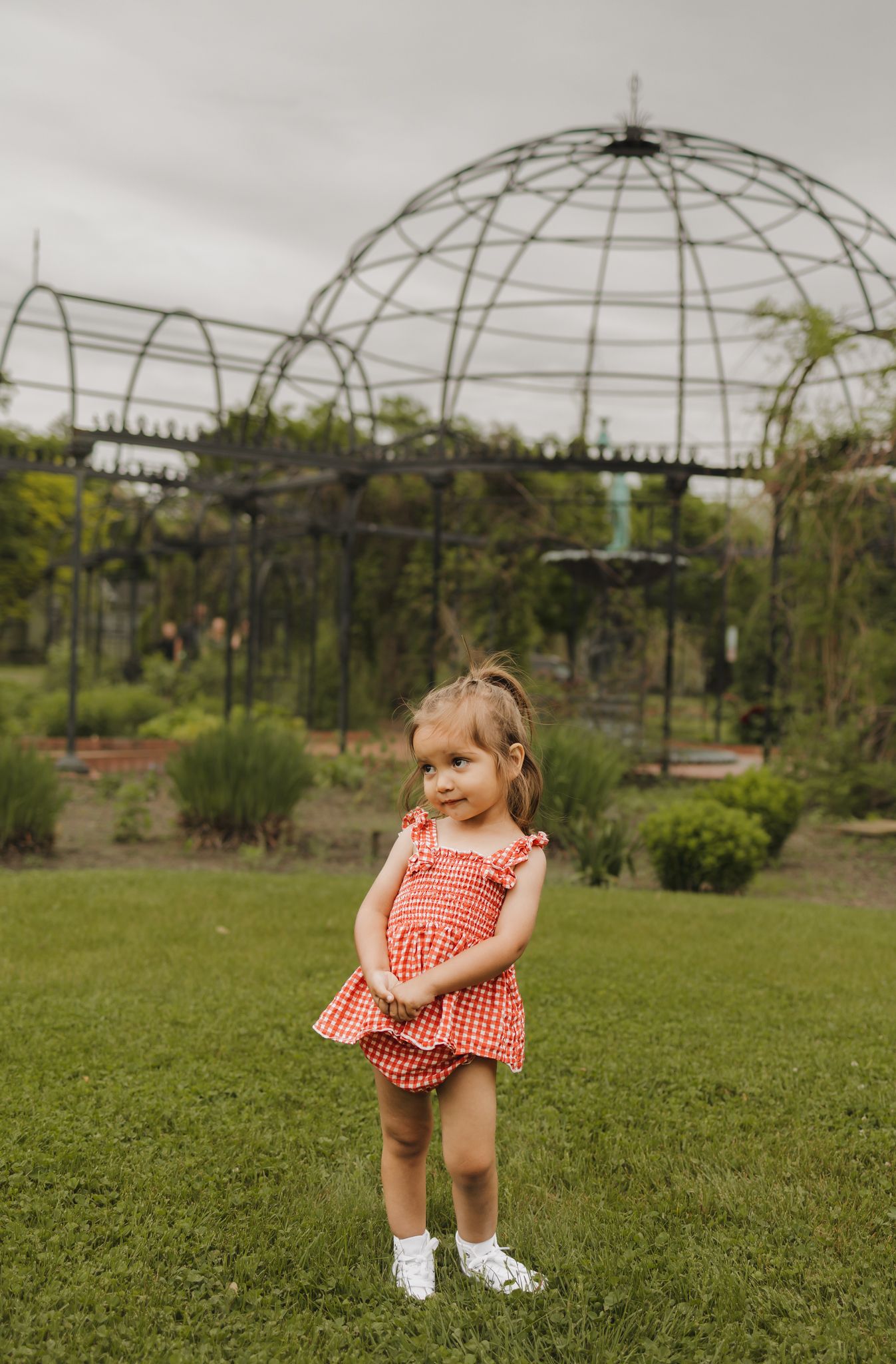 Toddler girl in red gingham dress stands on grass before ornate black metal garden gazebo.