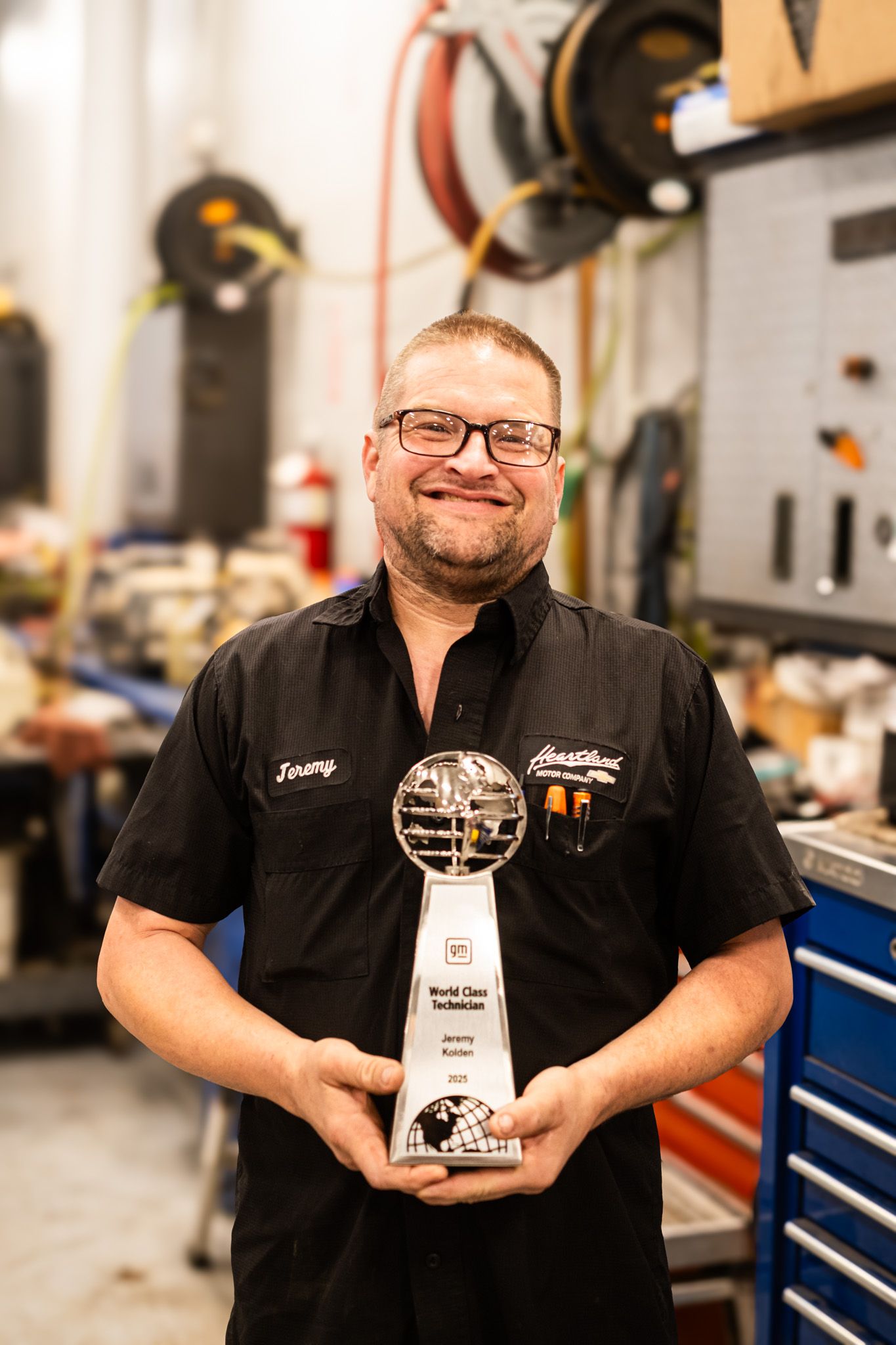 Auto technician holding World Class Technician trophy award in repair shop setting.