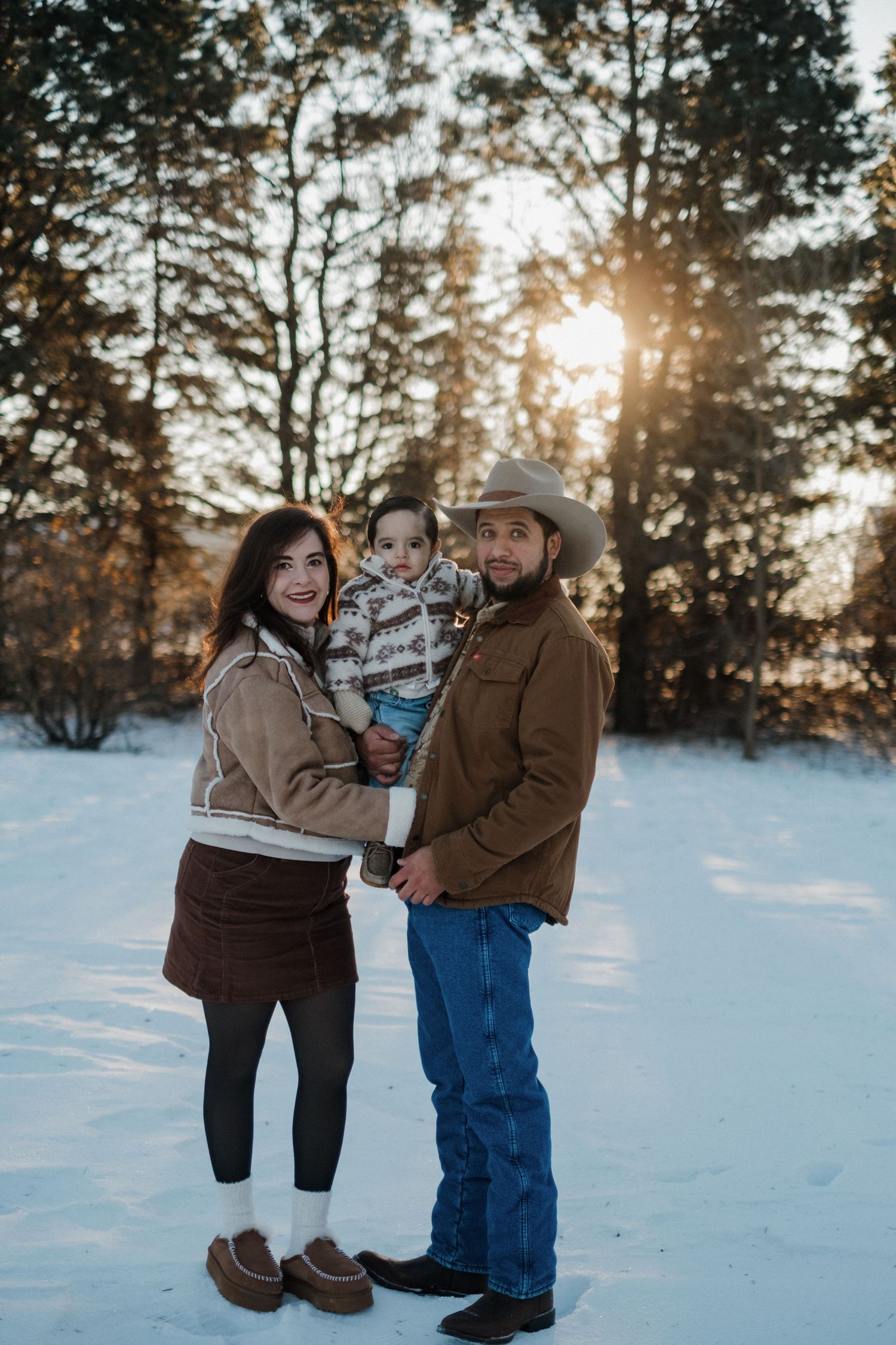 Family enjoying a winter photoshoot in the snow with evergreen trees and sunset in background.