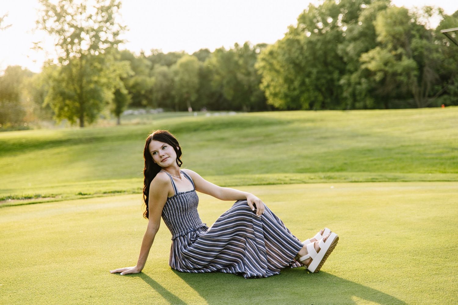 Young woman in striped maxi dress sitting on lush green golf course grass, smiling in golden hour sunlight.