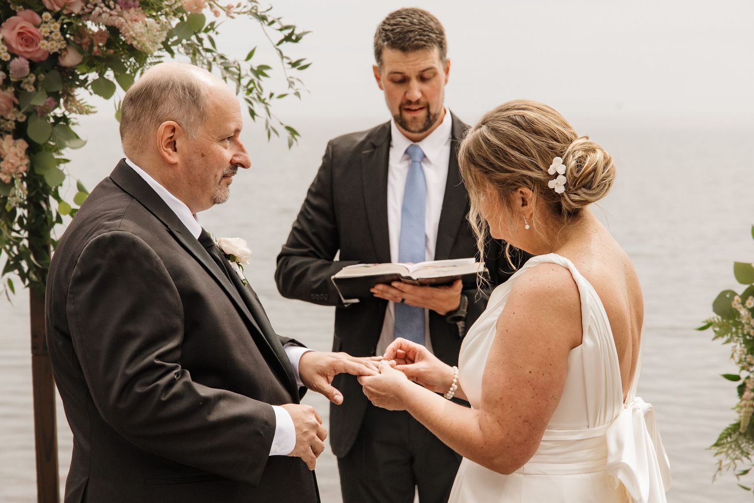 Bride placing wedding ring on grooms finger during outdoor ceremony with officiant and floral arch by the water.