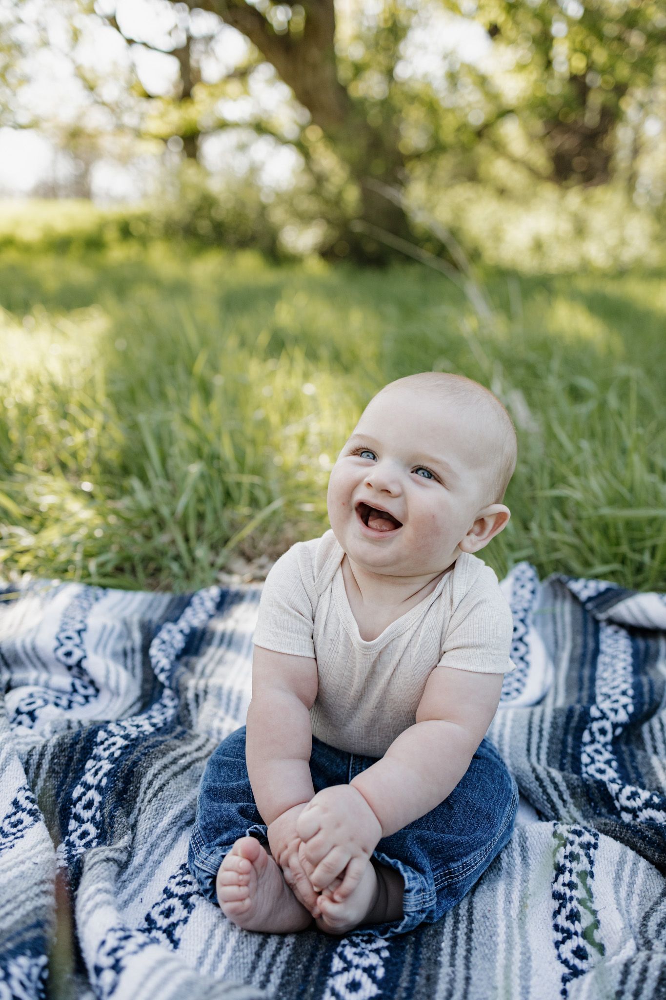 Laughing baby sitting on a patterned blanket outdoors in green grass, wearing a cream onesie and jeans.