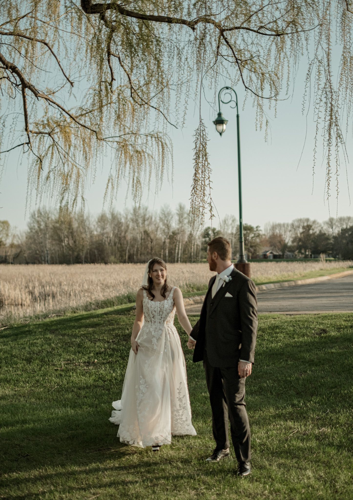 Newlyweds walking together on grassy hill with willow trees in background.