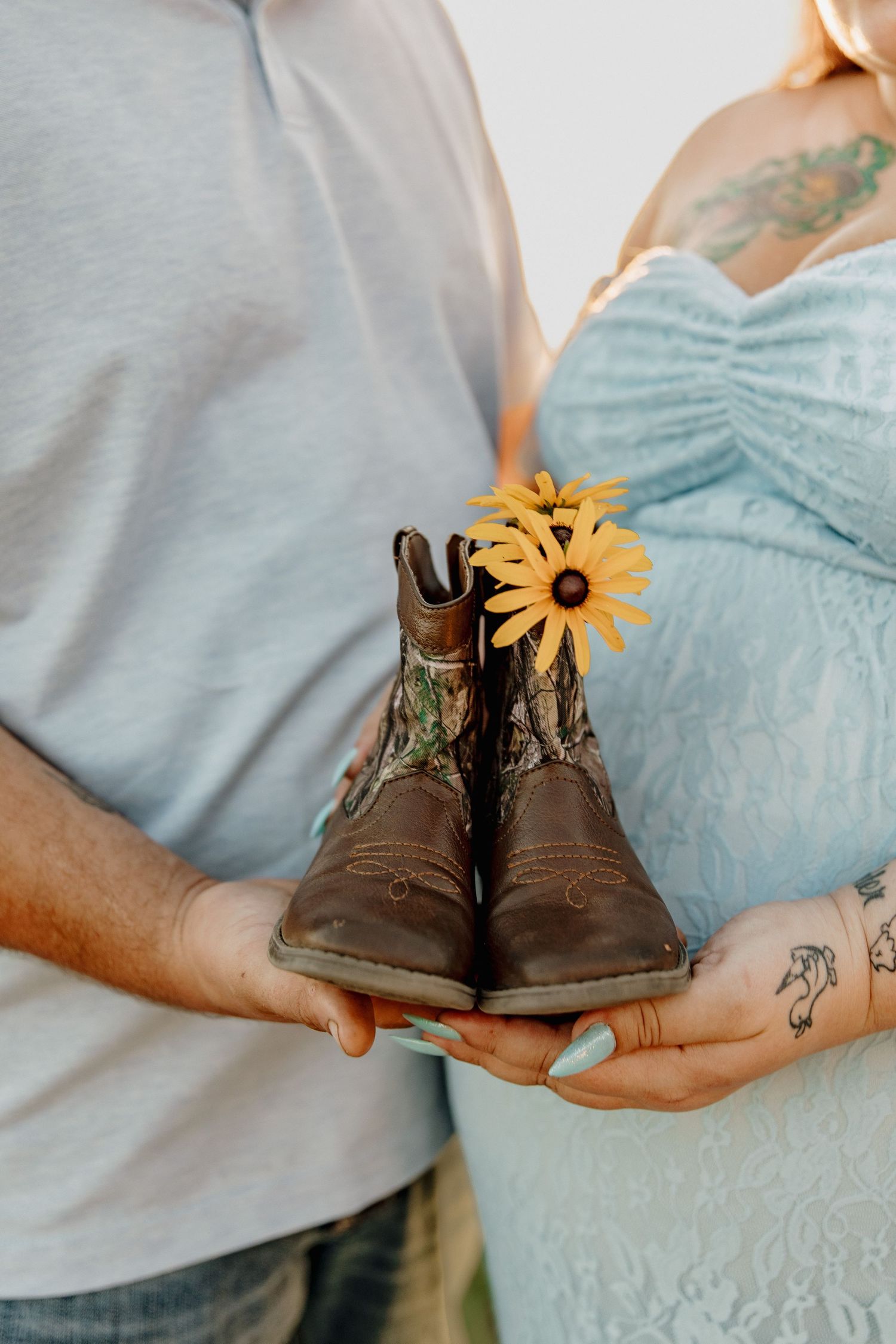 Pregnant couple holding tiny cowboy boots with yellow sunflowers, maternity announcement photo with tattoos visible.