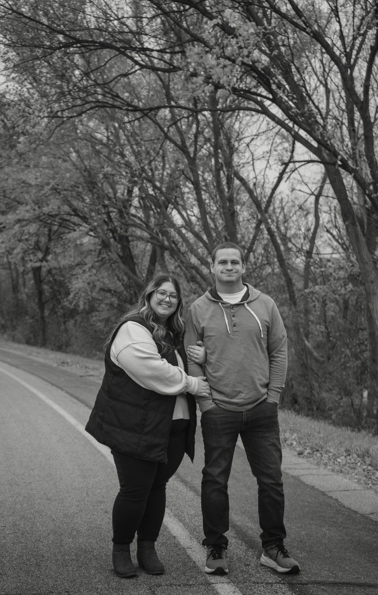 Black and white photo of a smiling couple standing together on a tree-lined road in autumn.