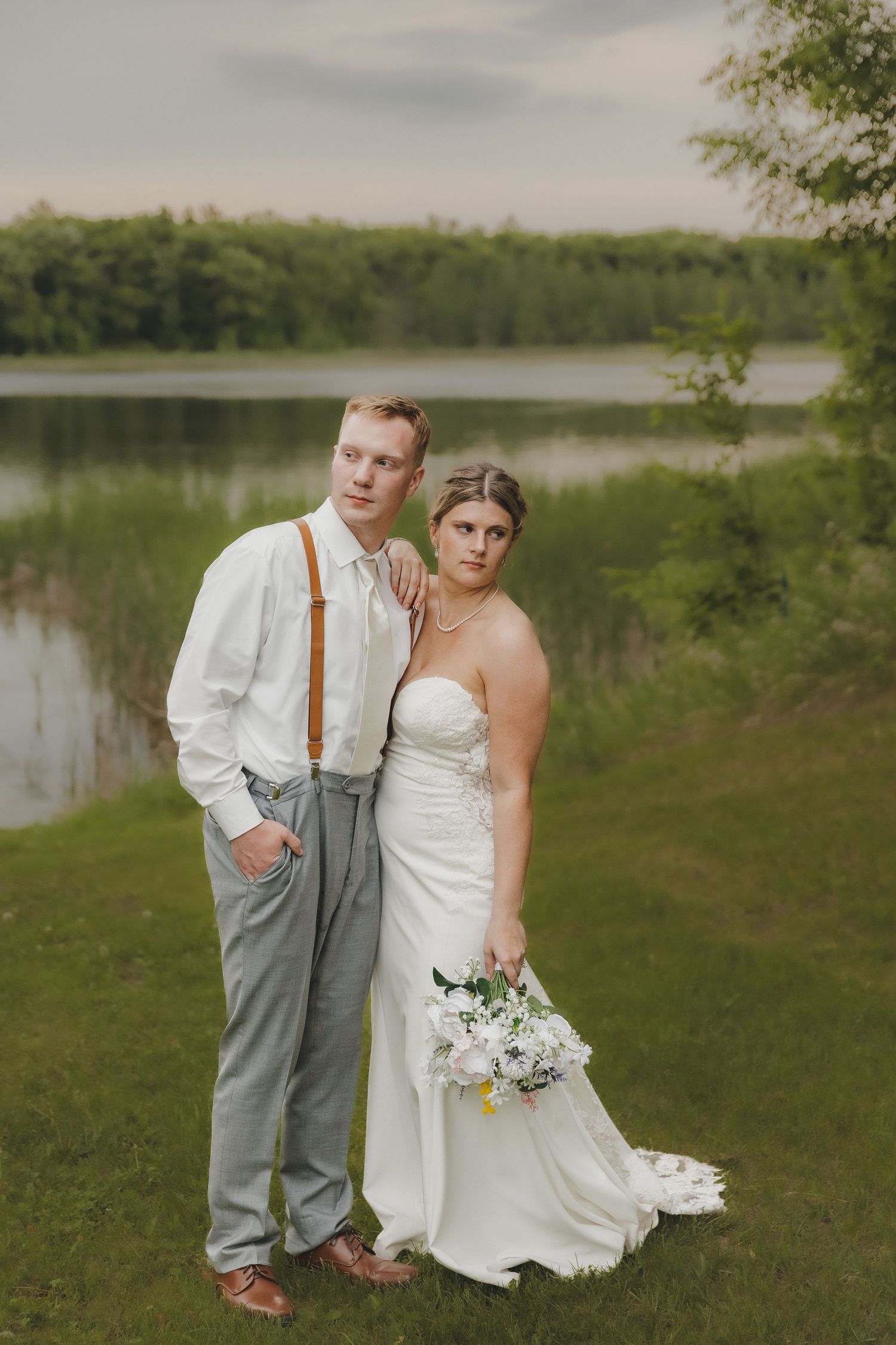 Bride and groom pose by serene lake, groom in white shirt with brown suspenders, bride holding white floral bouquet.