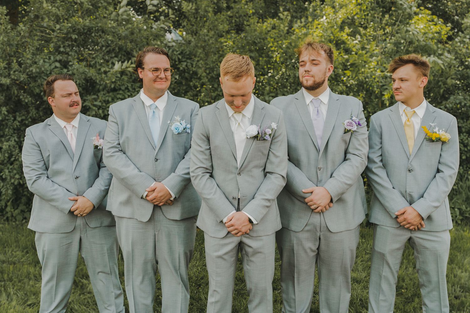 Five groomsmen in light gray suits standing outdoors, groom distinguished by boutonniere at wedding.