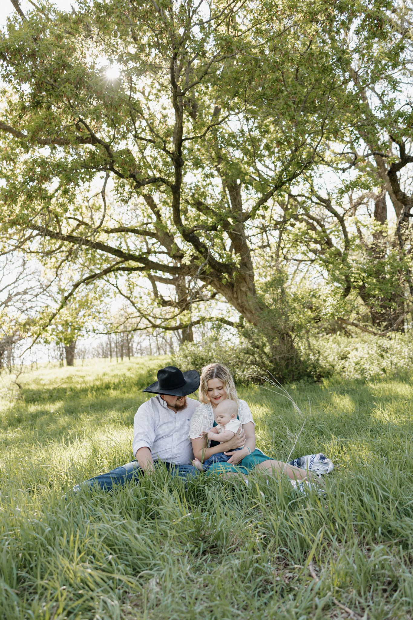 Family sitting on grass under a large oak tree, parents with baby enjoying a sunny outdoor picnic.