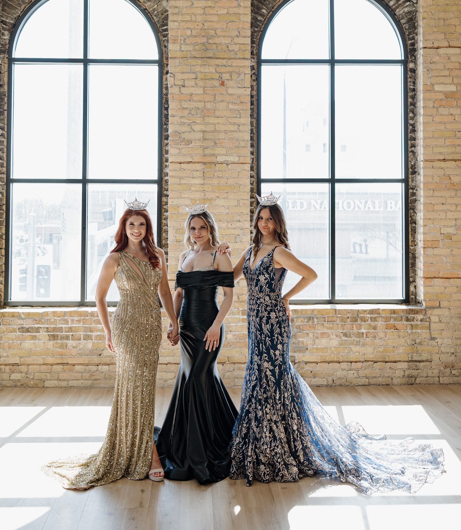 Three women in elegant gowns gold black and navy pose together in brick loft.
