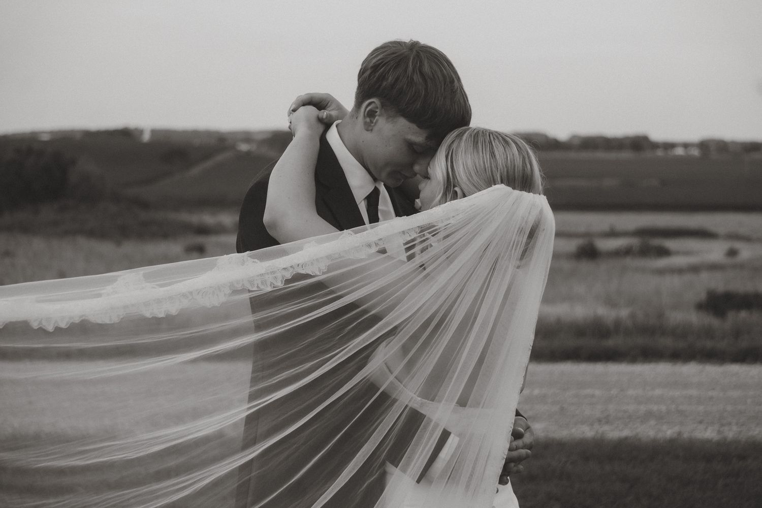 Black and white photo of a bride and groom sharing a romantic kiss, with her veil flowing in the wind at an outdoor wedding.