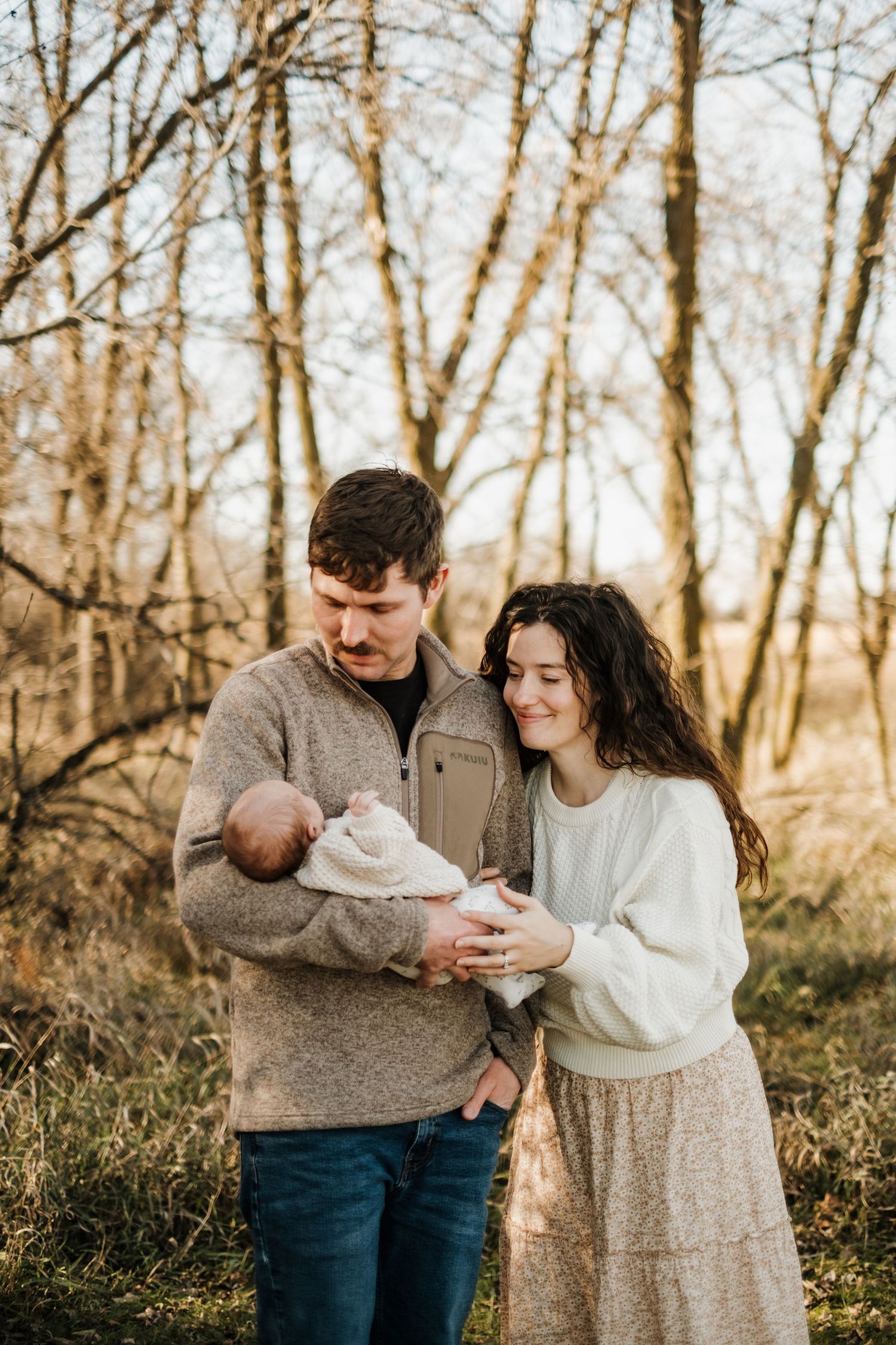 A couple shares an intimate moment with their newborn baby in a wooded setting during golden hour.
