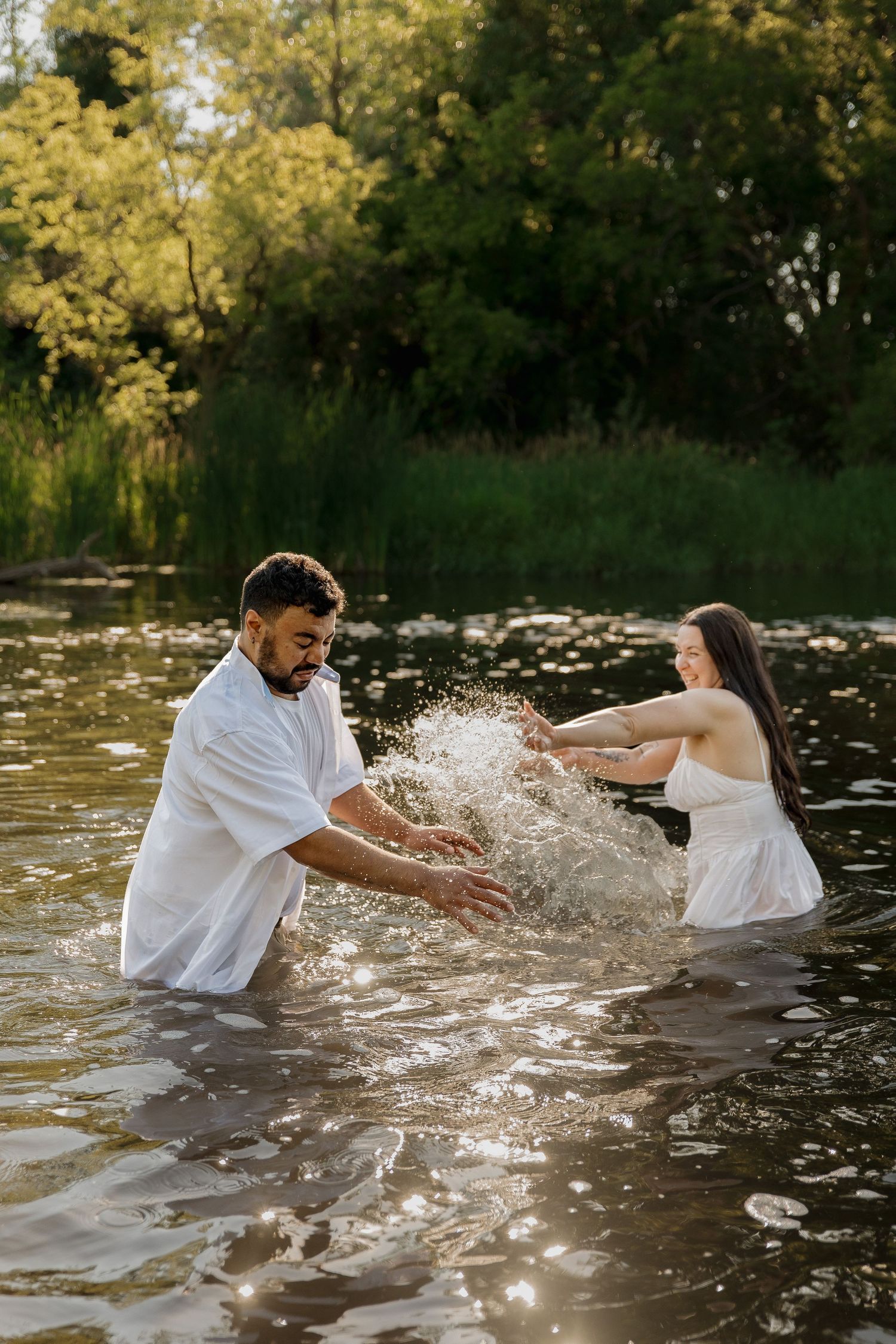 Couple in white outfits splashing water playfully in a sunlit river surrounded by lush green trees.
