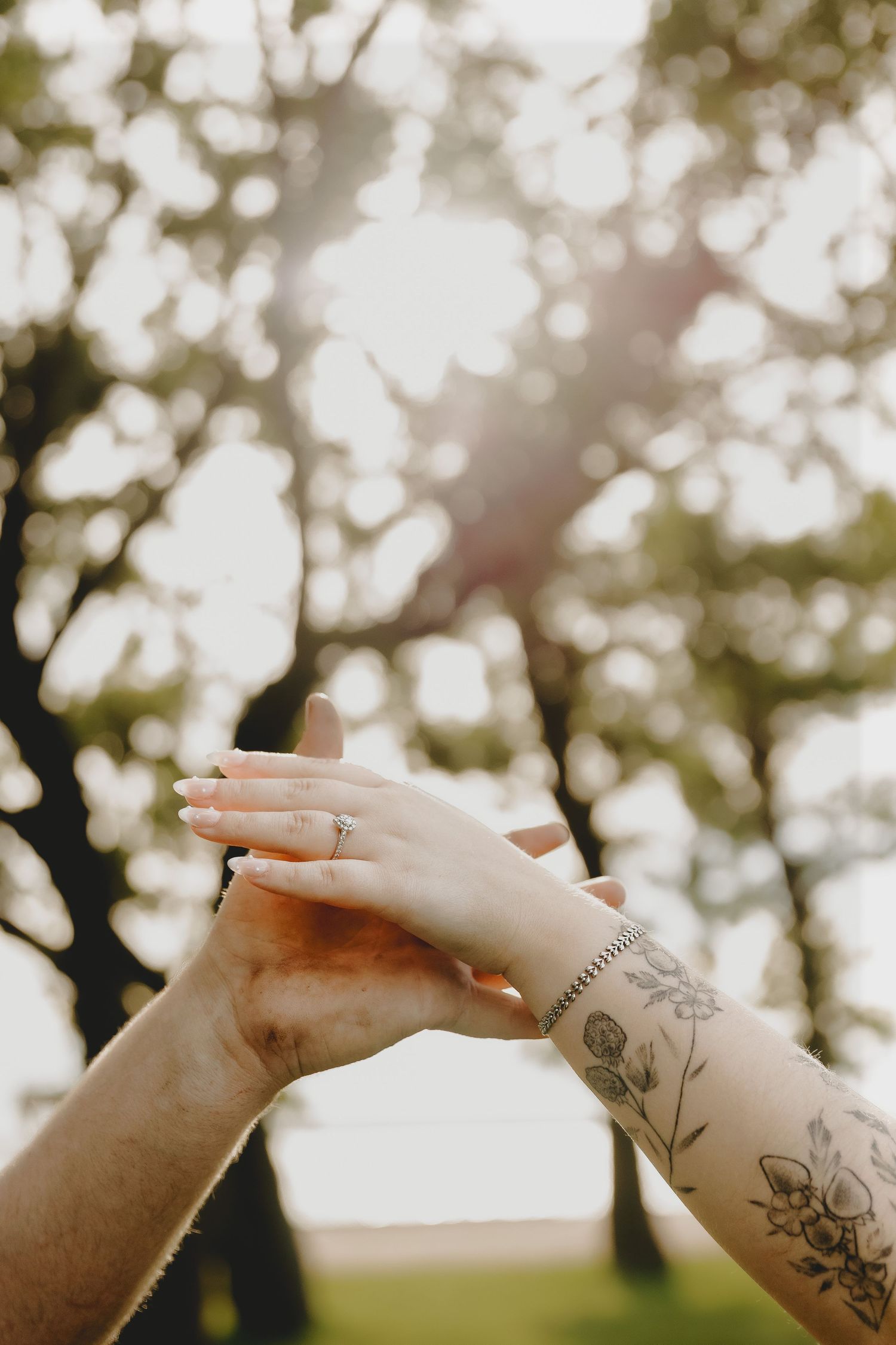 Close-up of couple's hands raised together, woman wearing engagement ring with floral tattoo on wrist, sunlit trees behind.