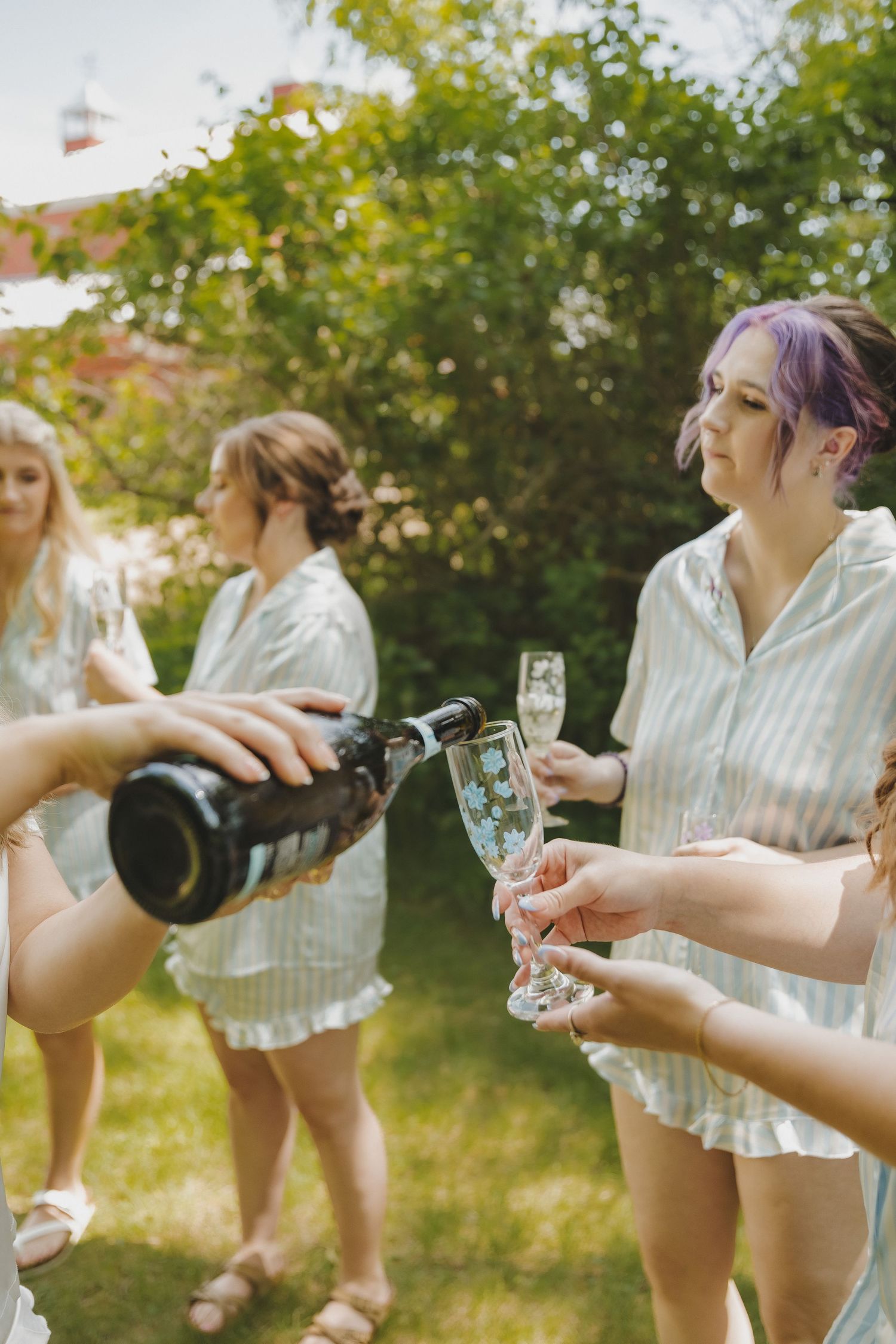 Bridesmaids in matching striped pajamas celebrate outdoors as champagne is poured into flutes on a sunny day.