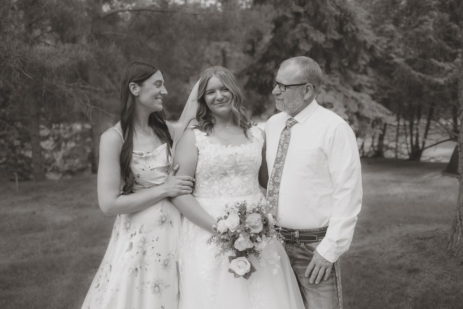 Black and white photo of smiling bride in wedding gown posing with two family members outdoors.