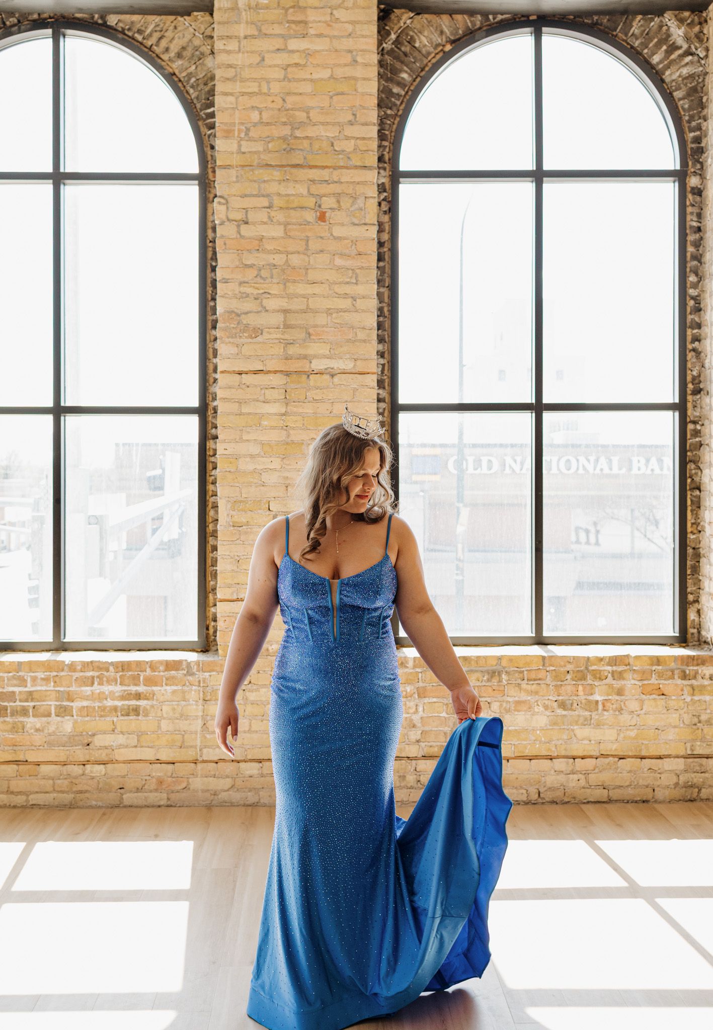 Woman in blue shimmer spaghetti strap mermaid gown poses by arched windows in brick loft.