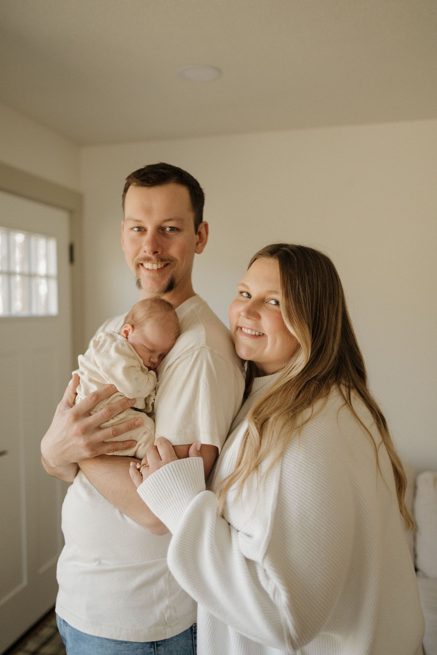 A smiling couple in white sweaters poses indoors, the man cradling a newborn baby in a bright, cozy room.