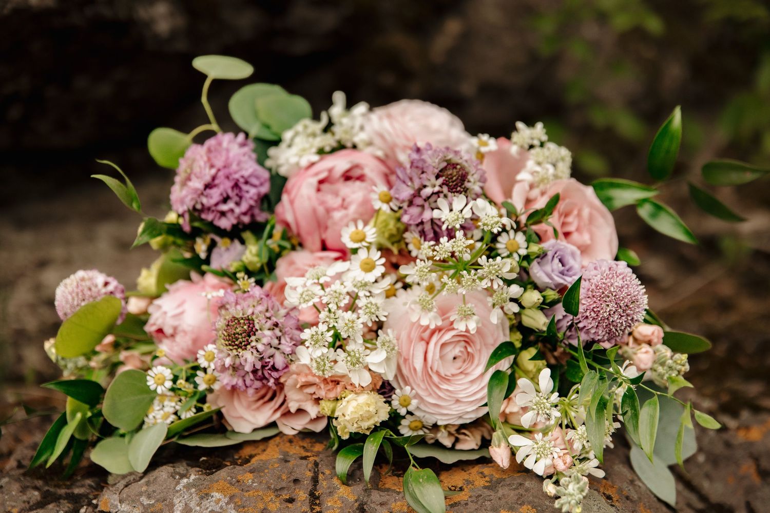 Romantic bridal bouquet with pink peonies, roses, dahlias, and white wildflowers on rustic stone surface.