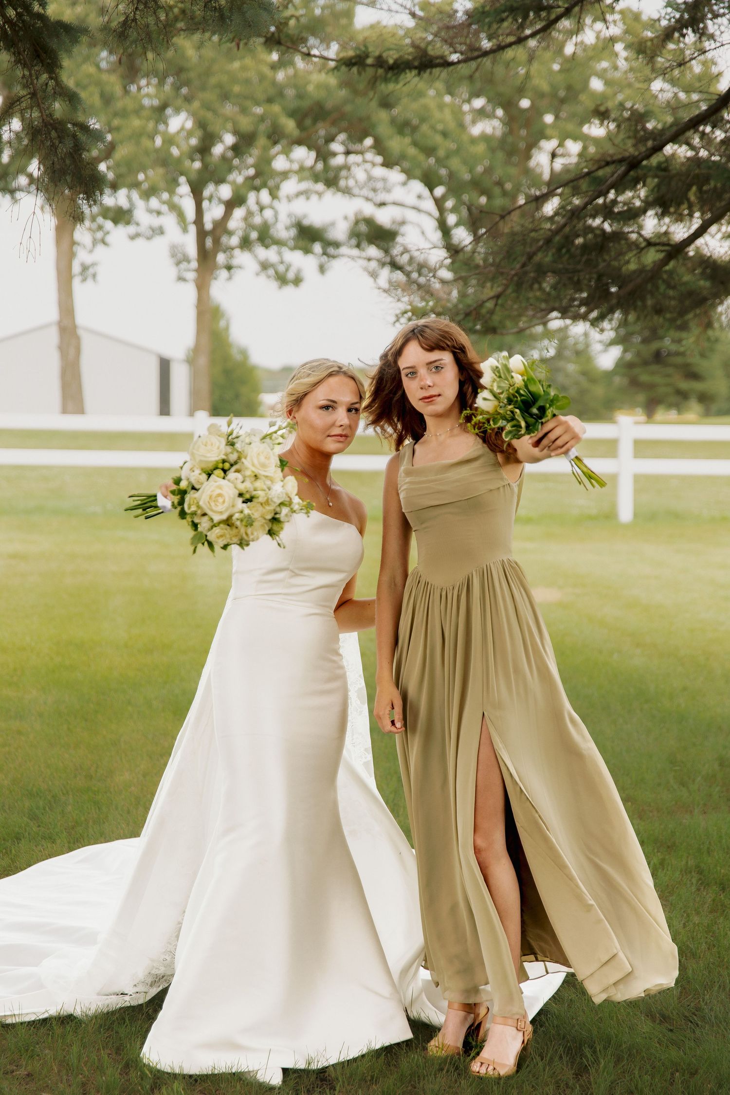 Bride in elegant white gown poses with bridesmaid in sage green dress holding white bouquets outdoors.