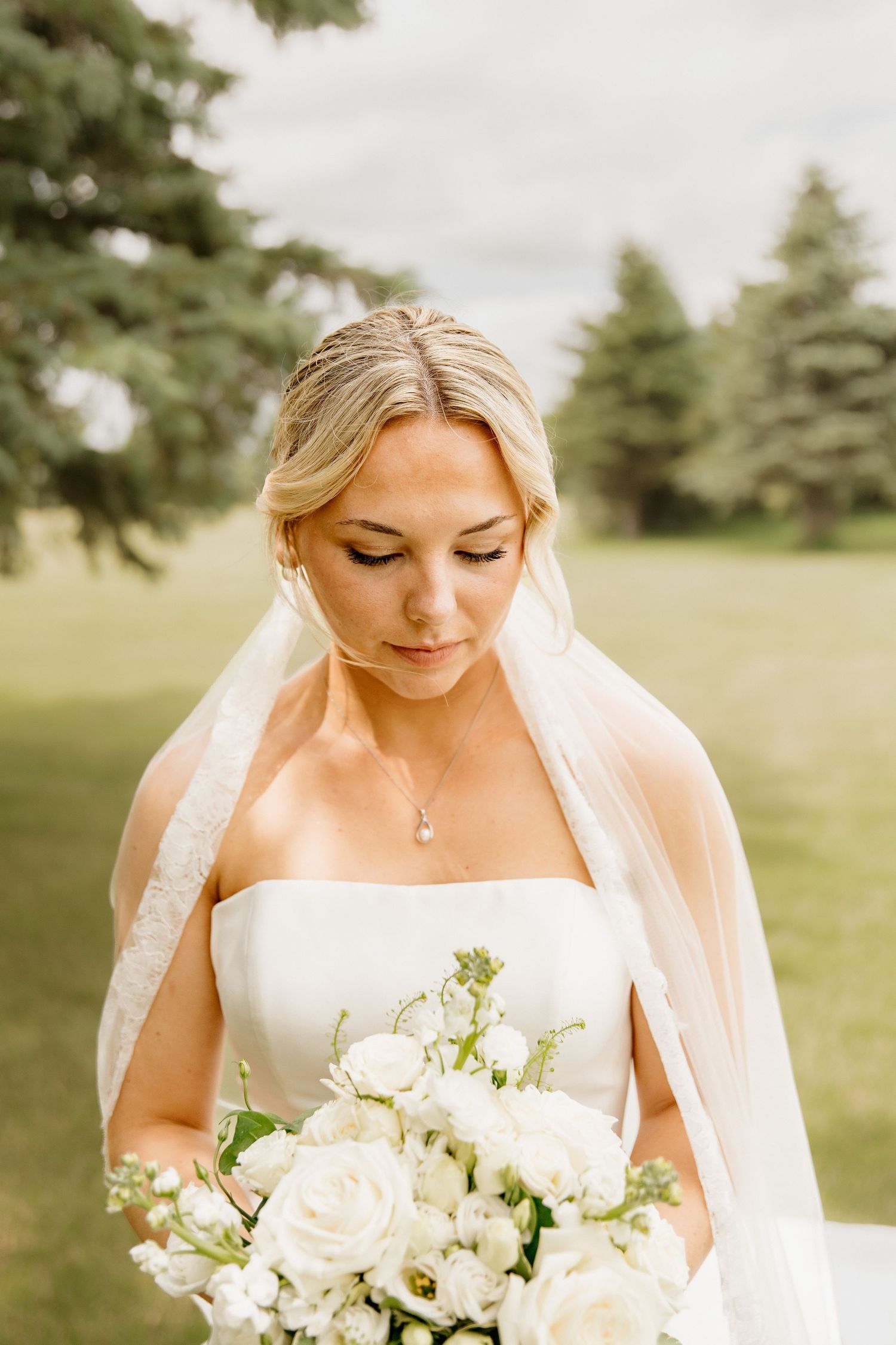 Blonde bride in strapless white gown holds white rose bouquet, wearing veil outdoors with green trees behind her.