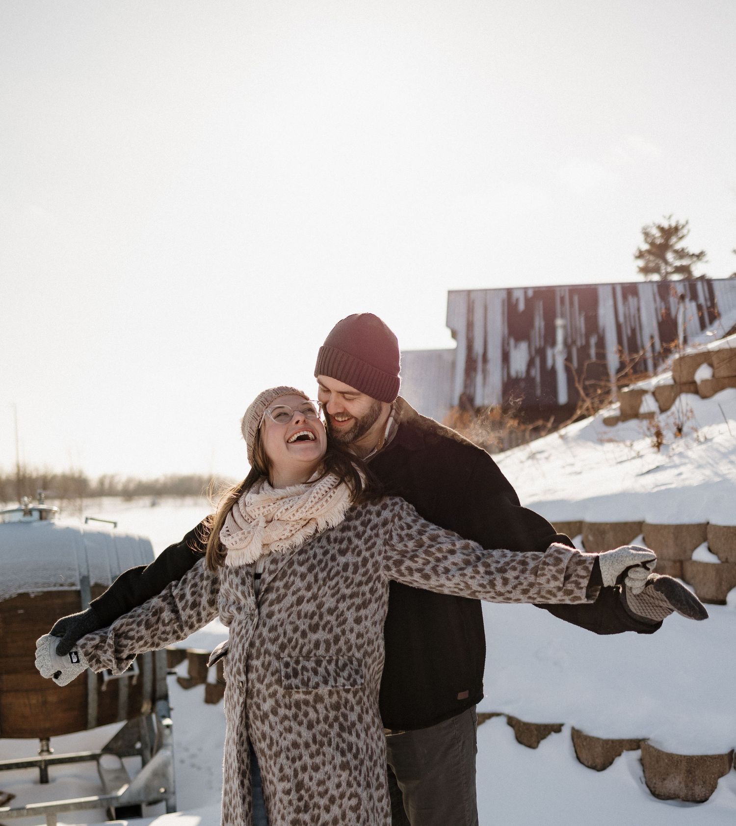 A couple in winter clothing embraces and laughs together in a snowy outdoor setting.