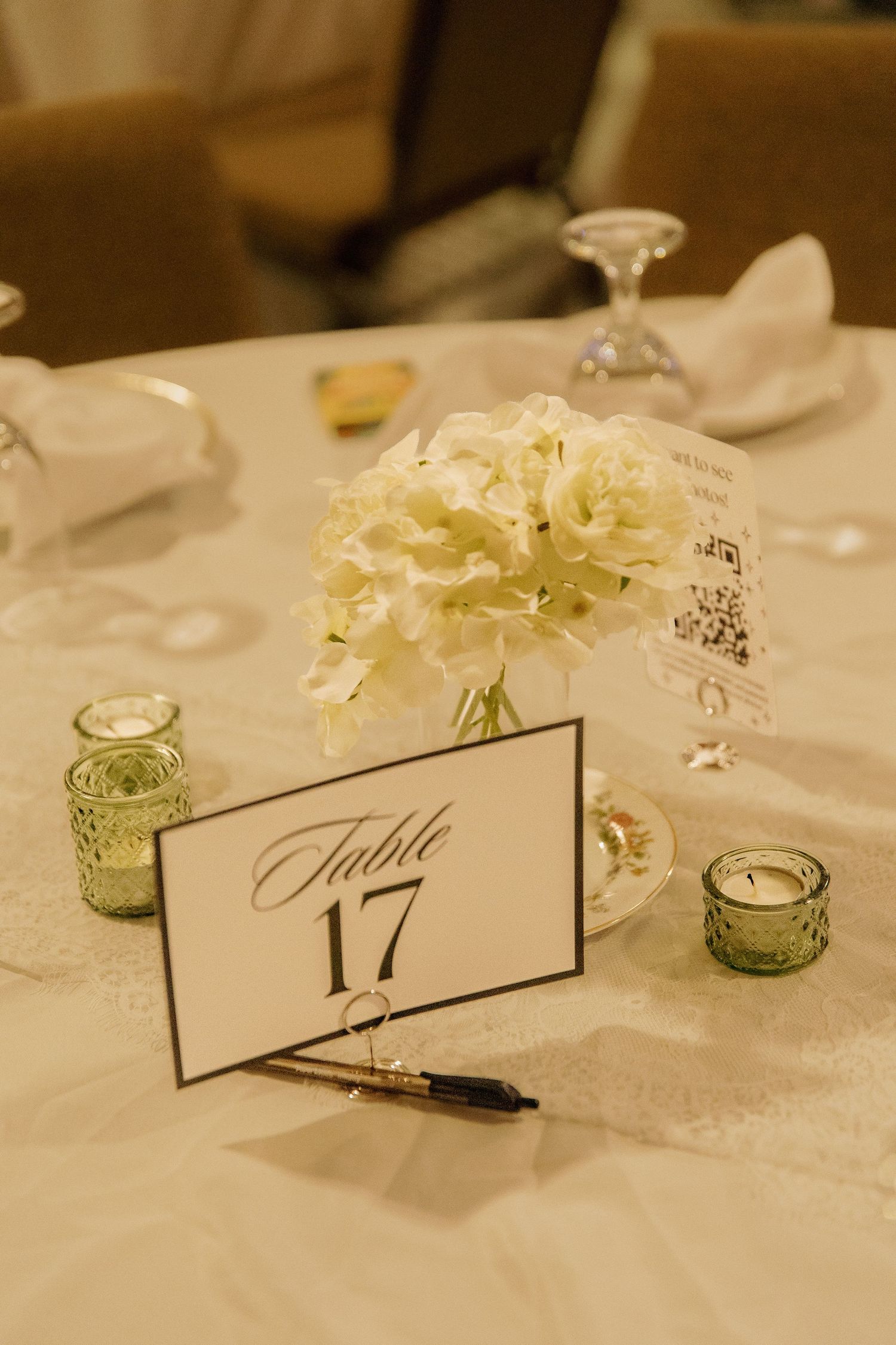 White floral centerpiece beside Table 17 sign on elegant white linen reception table with green votives.