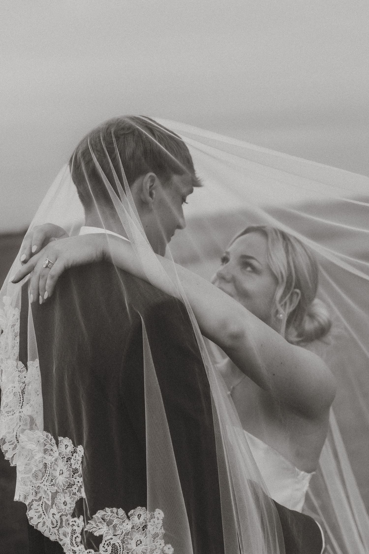 Bride and groom sharing romantic moment under lace-trimmed wedding veil in black and white.